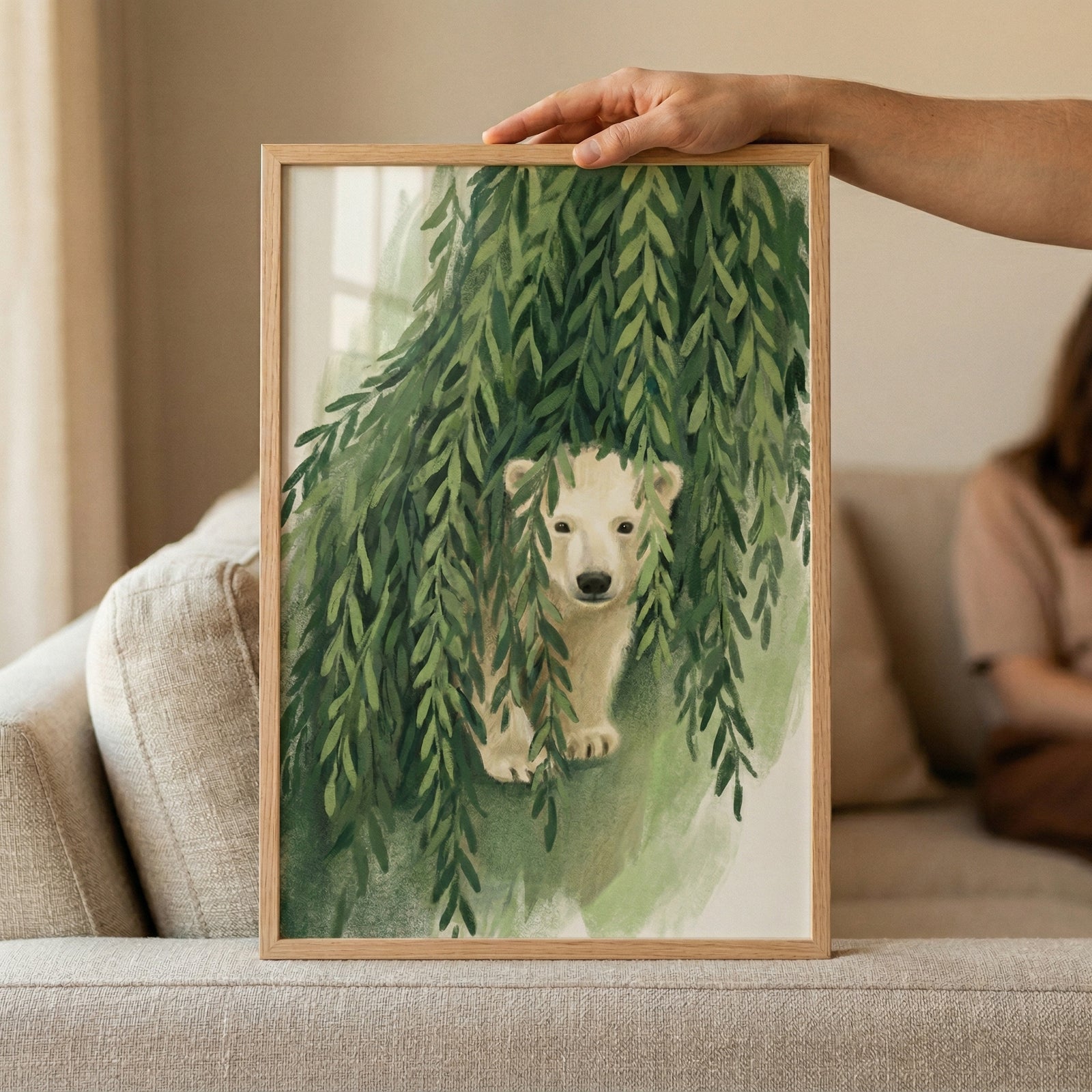 An illustration of a young white bear cub is featured within a light-colored wooden frame, which is being held by a hand. The cub is depicted peeking out from behind a dense thicket of long, green leaves that drape down like willow branches. The artwork has a soft, textured style with visible brushwork and a gentle color palette consisting mainly of greens and creams. The framed piece is positioned on a neutral-toned fabric sofa in a well-lit room.
