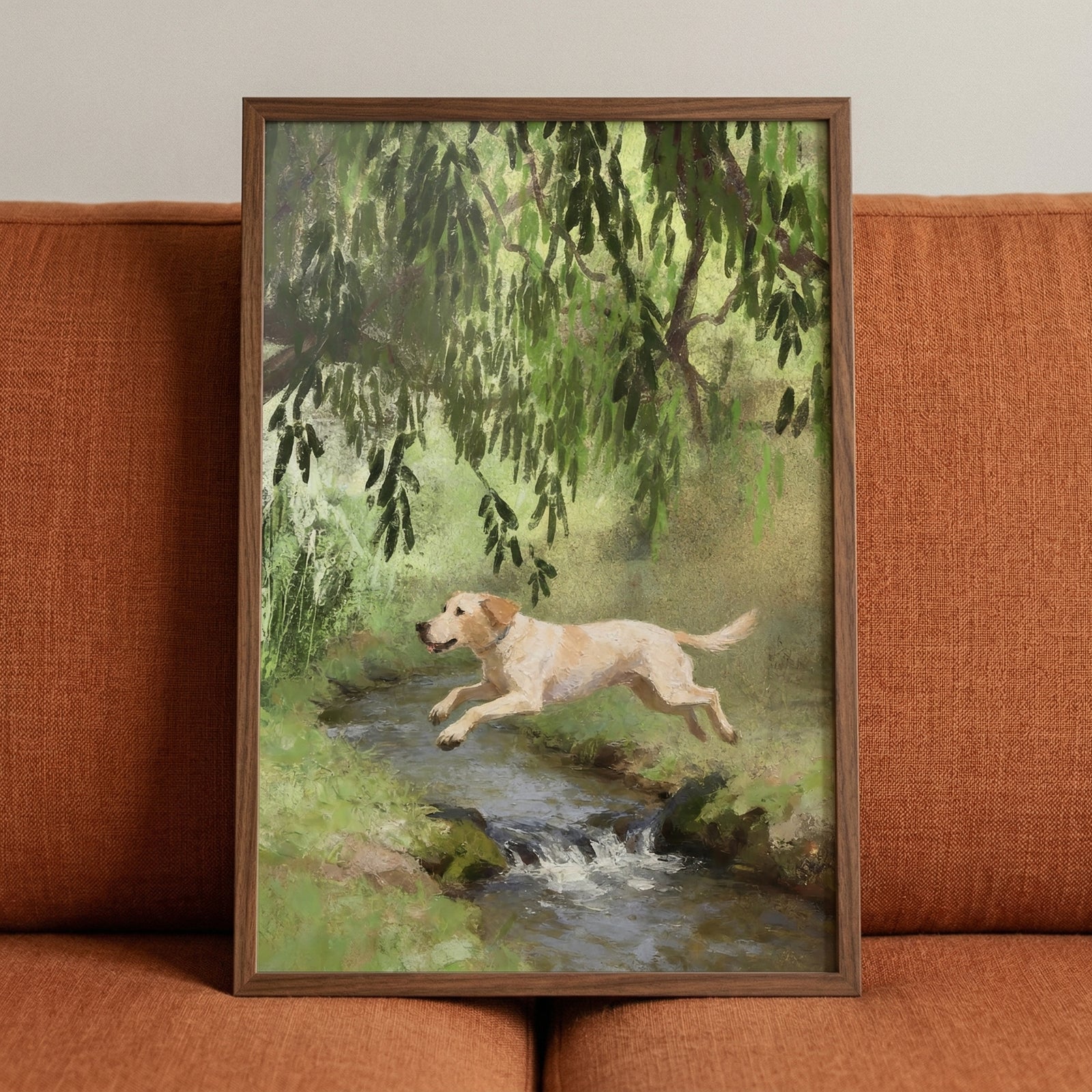 Framed painting of a yellow labrador mid leap over a small stream surrounded by lush green foliage and hanging tree branches resting against a rust colored sofa. The artwork captures motion and playfulness adding a lively natural focal point to the space.