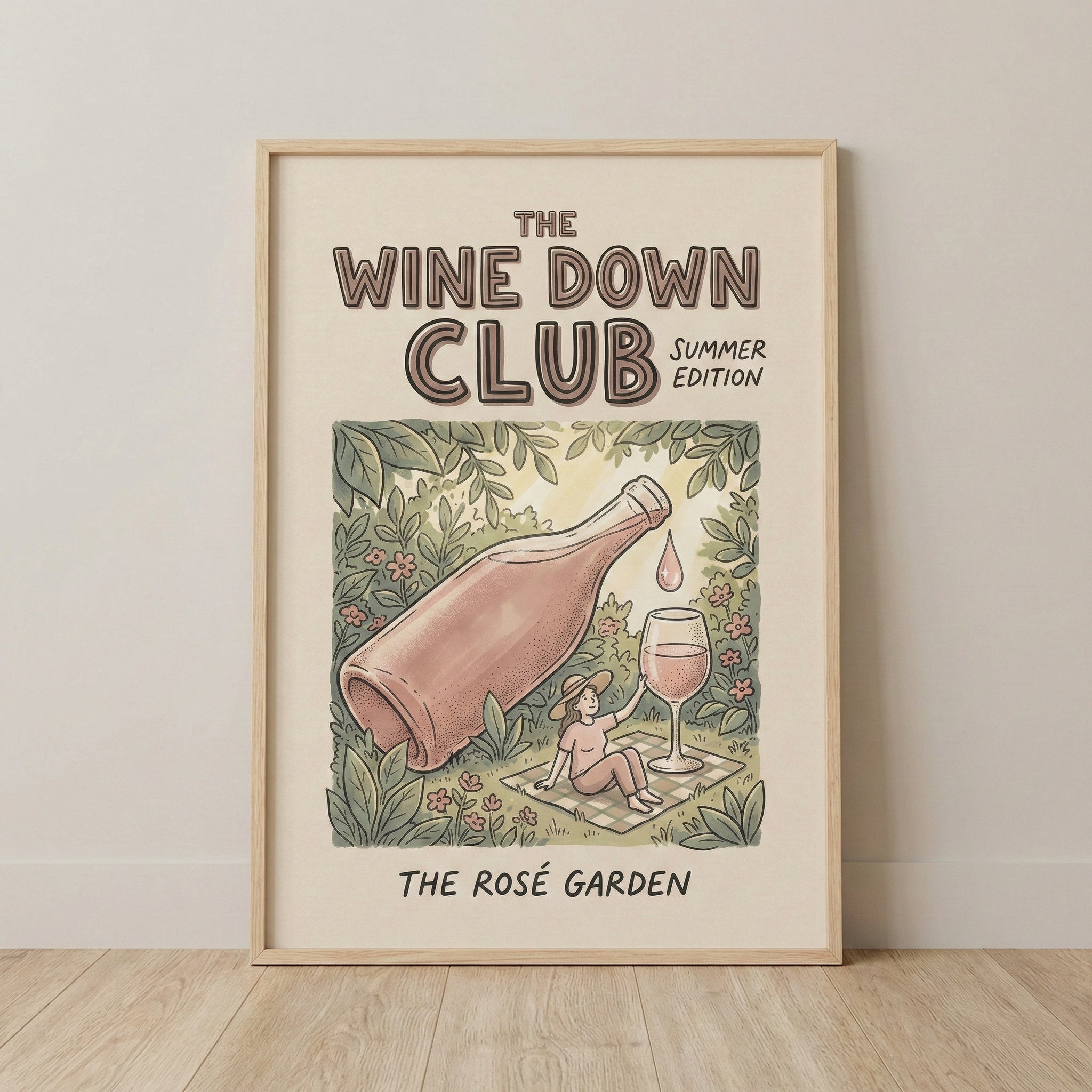 A framed poster titled “The Wine Down Club Summer Edition” shows an illustrated scene of a giant rosé bottle pouring a single drop toward a wine glass beside a person relaxing on a picnic blanket in a lush garden filled with flowers and greenery. The bottom text reads “The Rosé Garden,” and the artwork leans against a light wall on a wooden floor, giving the scene a soft, summery feel.