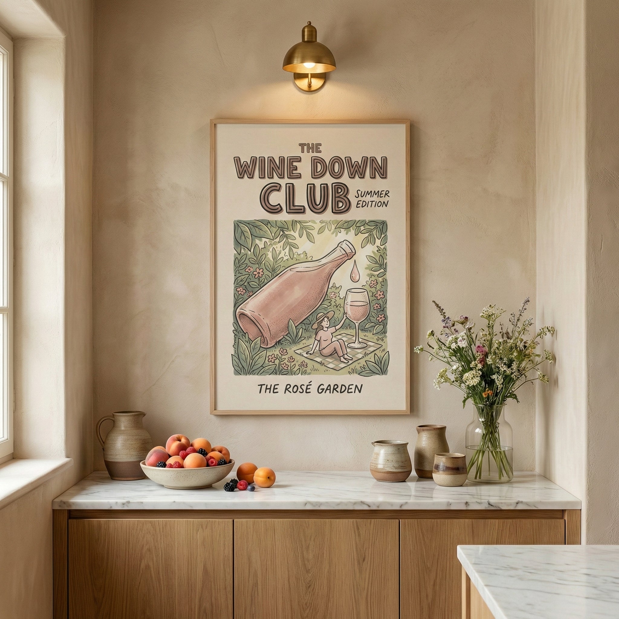 A framed poster titled “The Wine Down Club Summer Edition” hangs above a marble countertop, showing an illustrated scene of a giant rosé bottle pouring a drop toward a wine glass beside a person lounging on a blanket in a lush floral garden, with the text “The Rosé Garden” below. Fresh fruit, ceramic vessels, and a vase of wildflowers sit on the counter beneath warm wall lighting, creating a soft, summery kitchen nook.