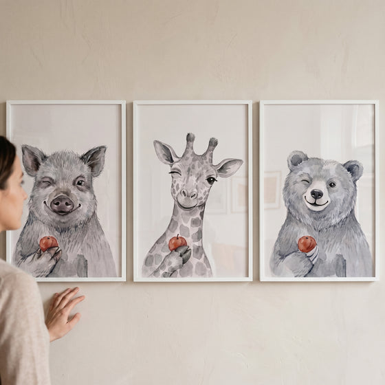 A person views a winking apple watercolor animals wall art set of 3 featuring a pig, giraffe, and bear.