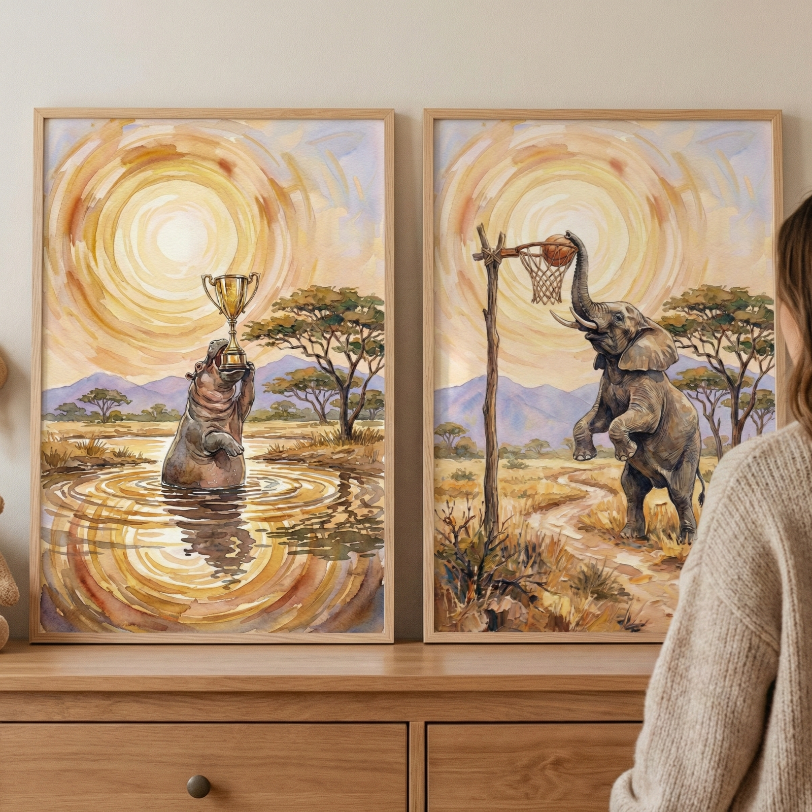 Two framed illustrations sit on a wooden dresser, depicting whimsical animal scenes in a savanna landscape. The left artwork features a hippopotamus in water, triumphantly holding a gold trophy. The right artwork shows an elephant on its hind legs, using its trunk to place a ball into a rustic basketball hoop. Both pieces share a warm color palette with a stylized, glowing sun and distant mountains under a golden sky. A person's shoulder is partially visible on the right side.