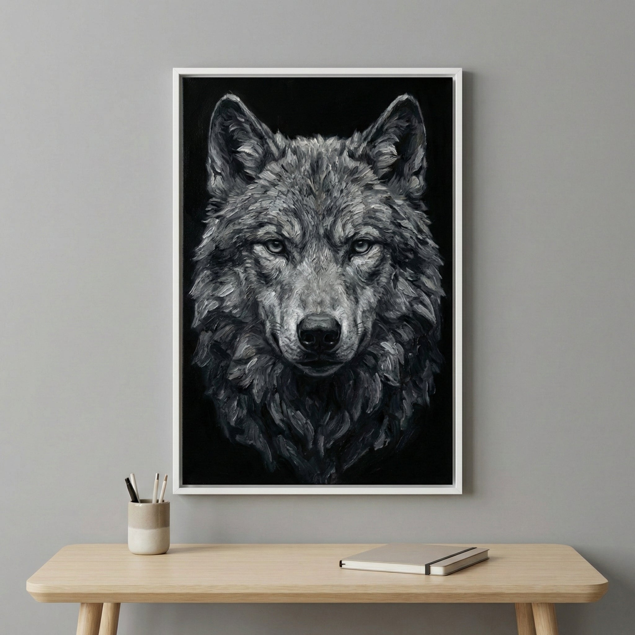 Framed painting of a wolf with intense, piercing eyes and textured gray fur, hung on a light gray wall above a minimalist wooden table with a cup of pens and a closed notebook. The wolf's detailed expression and dark background create a bold focal point.
