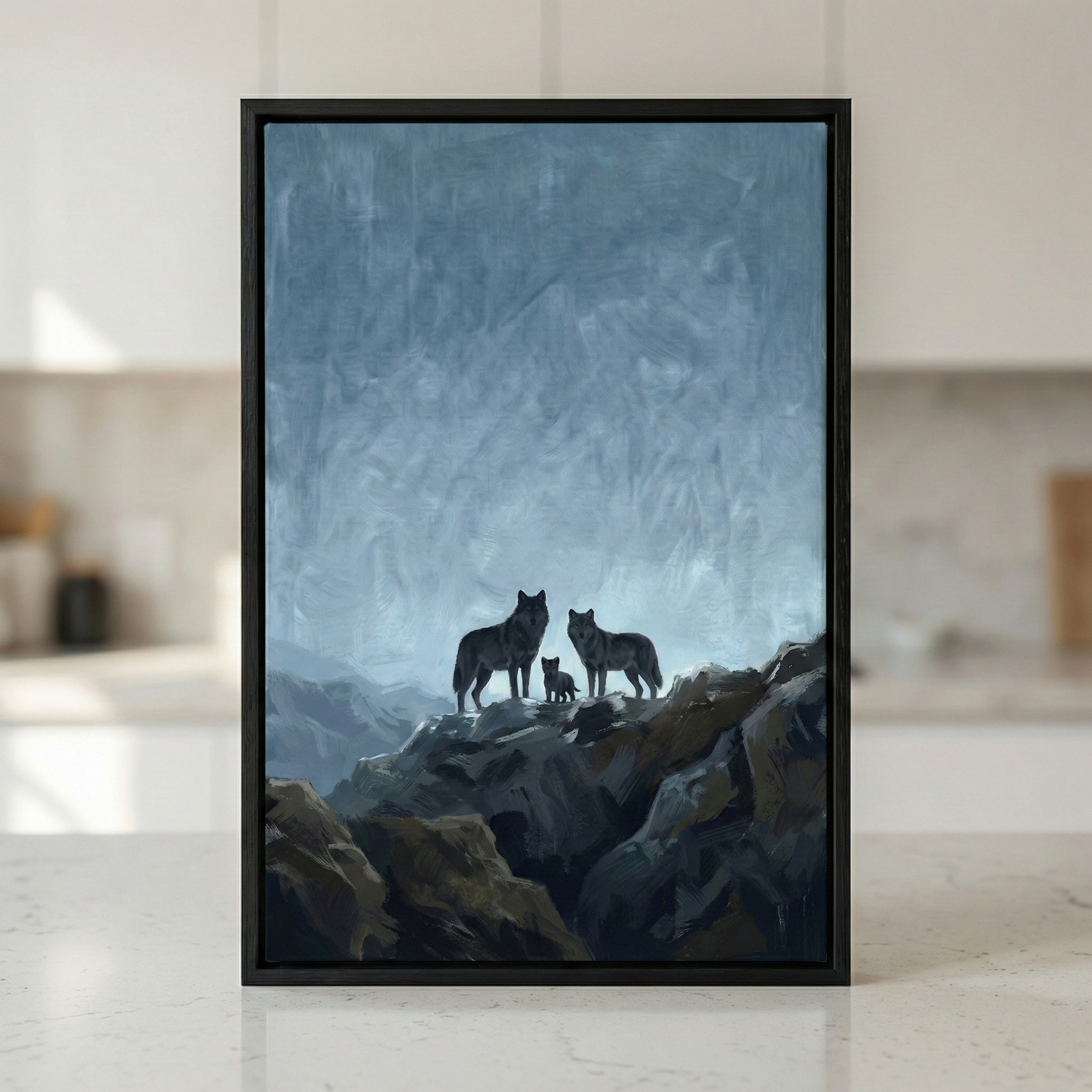 A framed piece of art depicts a wolf family standing on a rocky outcrop. Two adult wolves flank a small pup, silhouetted against a textured, pale blue and grey sky. The artwork features visible, broad brushstrokes that add texture to the atmospheric landscape and the dark, craggy mountain peak. The piece is set in a thin black frame and sits on a light-colored, speckled surface in a home environment.