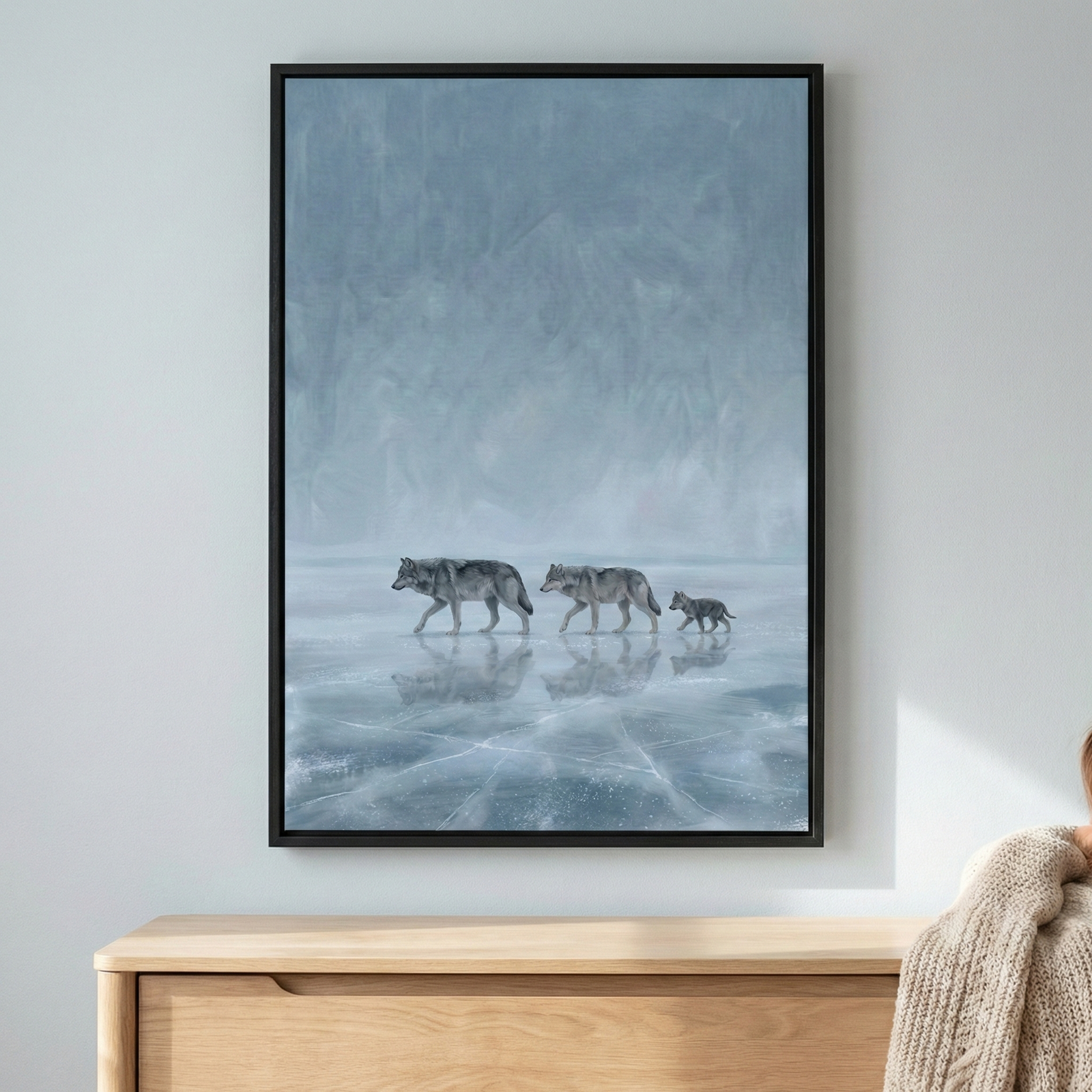 A vertical framed artwork hangs on a pale wall above a light-wood dresser. The piece depicts three wolves—two larger adults and one small pup—walking in a line across a frozen, cracked lake. Their forms are reflected on the icy surface below. The upper two-thirds of the image features a misty, textured background in shades of cool blue and grey, creating an atmospheric winter wilderness scene.