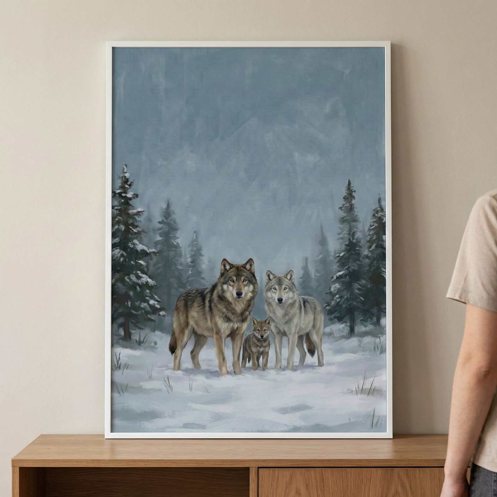 This vertical artwork depicts a family of three wolves in a winter setting. In the center, two adult wolves stand protectively near a small pup. Their thick fur is rendered in realistic shades of grey, brown, and white. They are set against a landscape of snowy ground, dark evergreen trees, and distant, misty mountains under a pale blue sky. The image features visible brushwork and a textured finish. It is displayed in a white frame on a wooden console against a neutral wall.