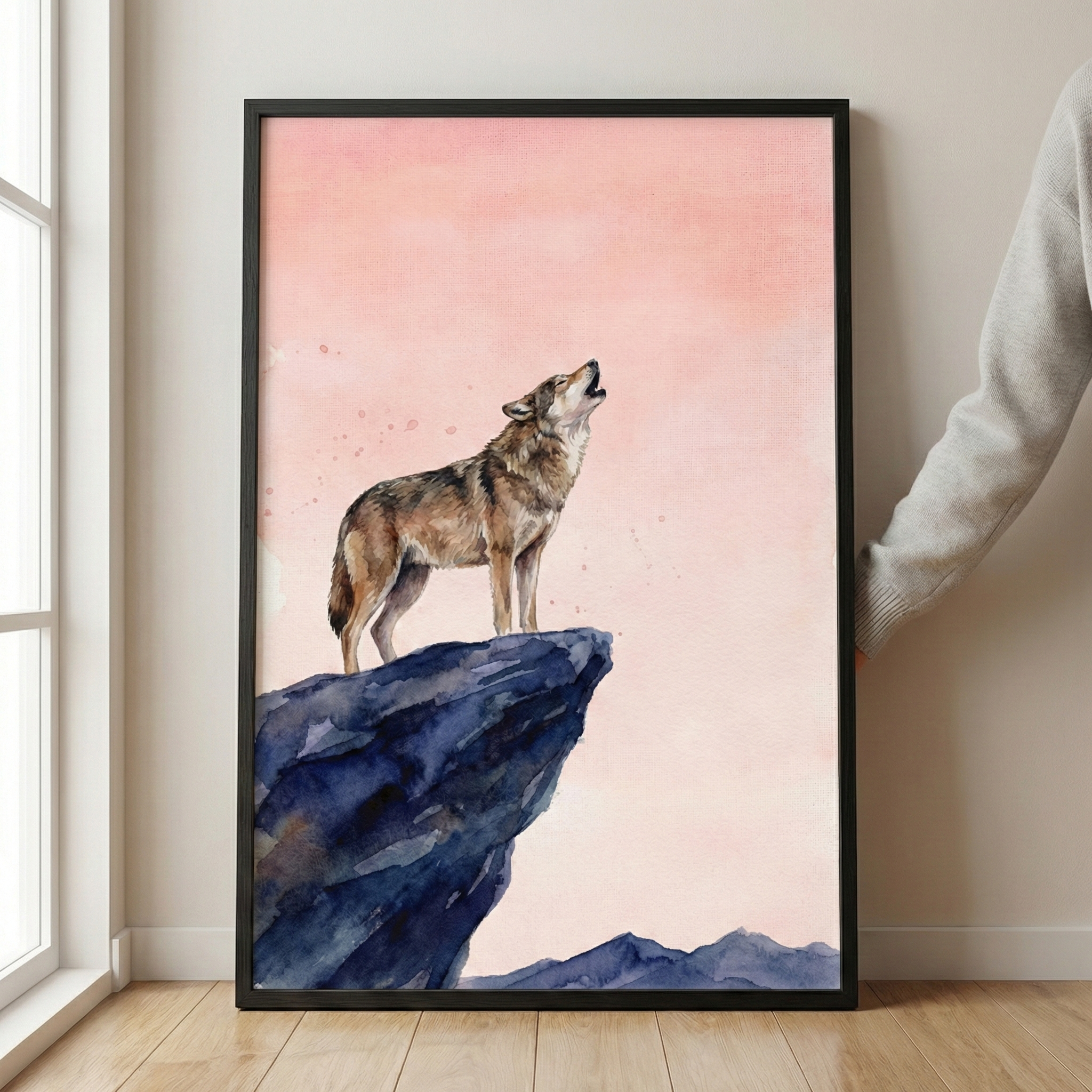 A framed watercolor illustration shows a lone wolf howling from the peak of a dark blue, craggy rock. The background is a soft gradient of pale pink and peach tones with subtle watercolor textures. The wolf is detailed with earthy shades of brown and grey. The piece is set in a black frame, resting against a minimalist light wall and a natural wood floor. A person's arm in a grey sleeve is seen holding the side of the frame.