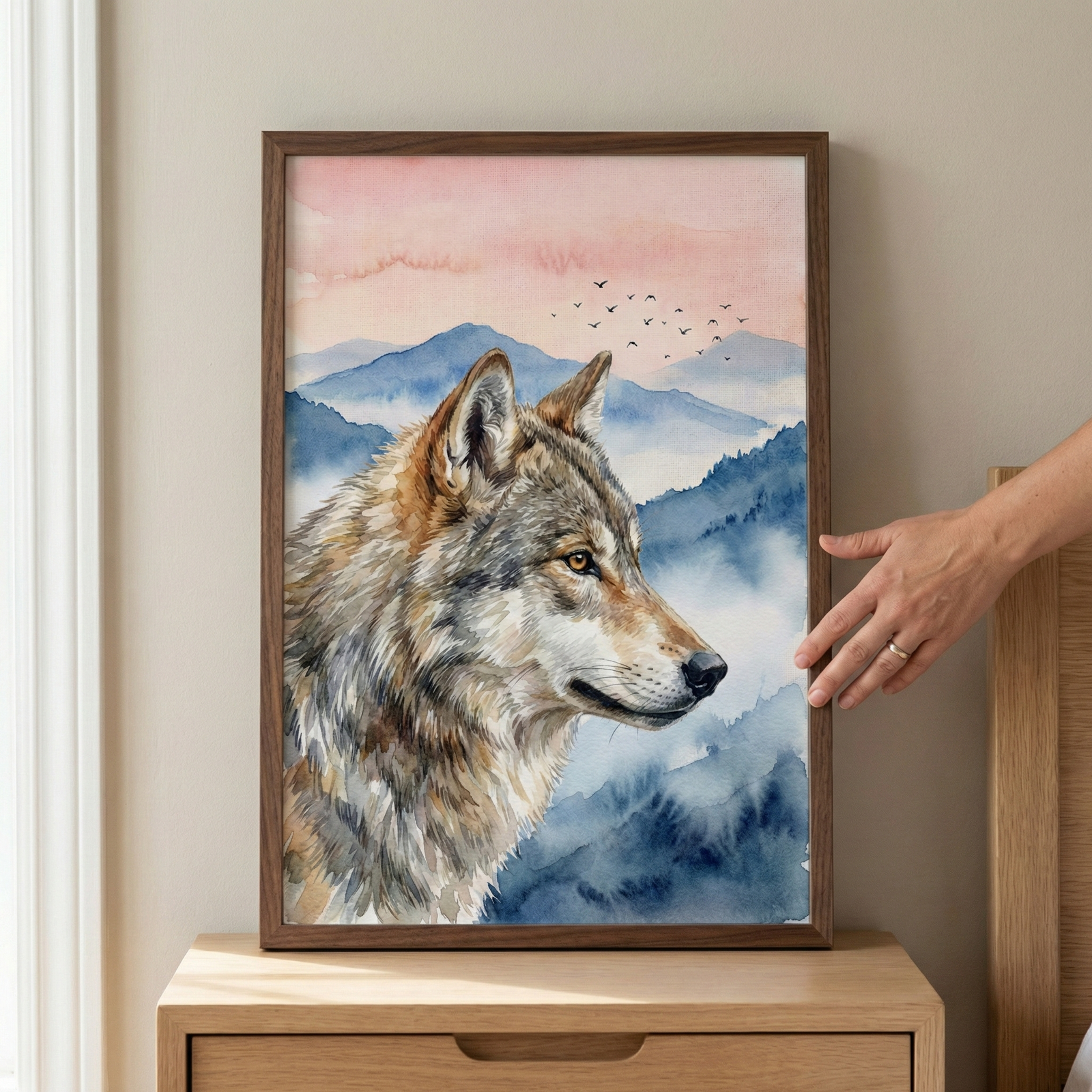 A framed watercolor-style illustration of a wolf's head in profile. The wolf has detailed fur in shades of grey, brown, and white. Behind the animal, soft blue mountain peaks recede into a hazy distance under a pale pink sky. A small flock of birds is silhouetted against the sky. The artwork is held in a slender dark wood frame, sitting on a light-colored wooden dresser. A person's hand is visible on the right side, touching the edge of the frame.