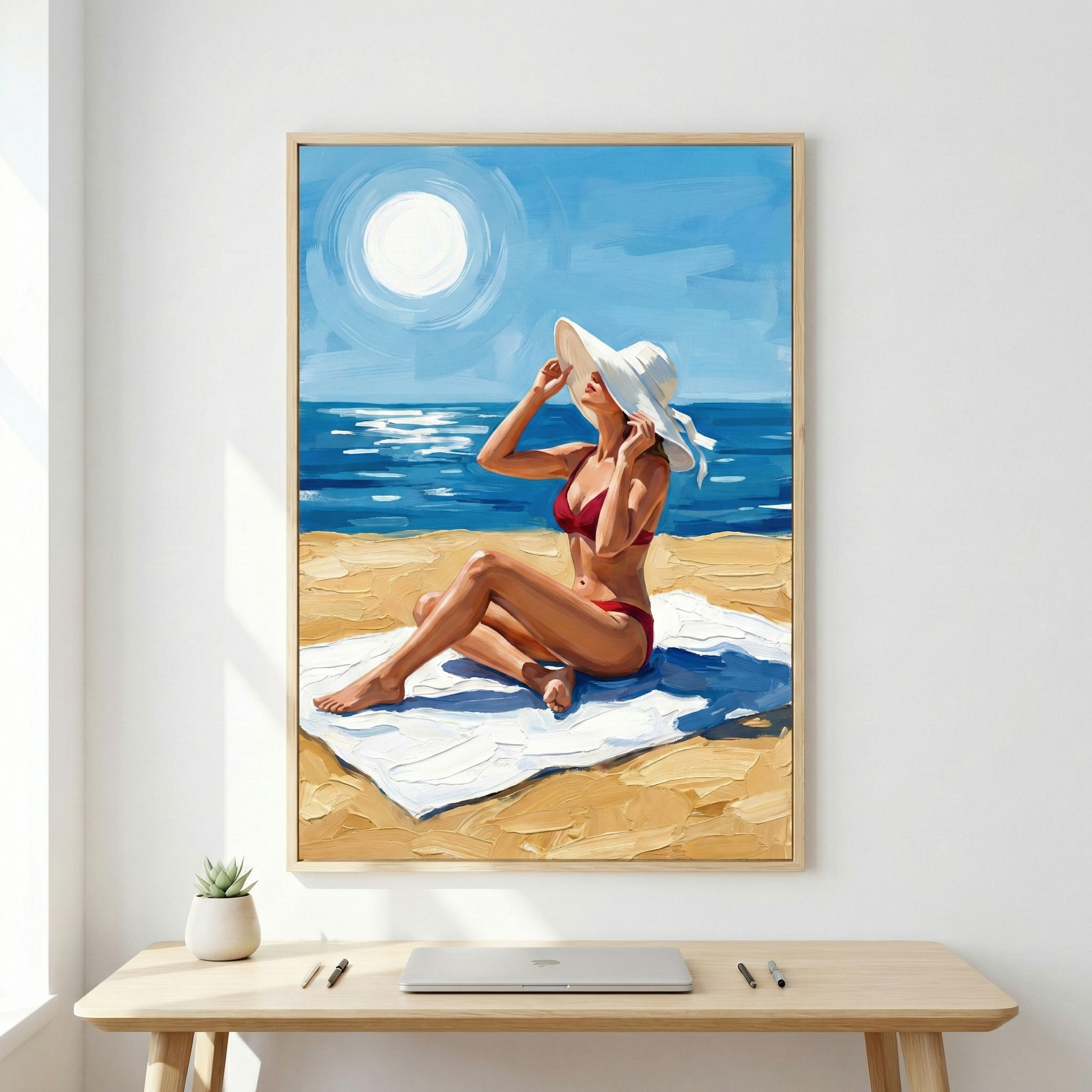 A framed artwork displays a woman in a red swimsuit and a large white sunhat, relaxing on a white towel on a sun-drenched beach. The scene shows a deep blue sea and a bright sky with a prominent, glowing sun, all rendered with thick, expressive strokes. The light wooden frame hangs on a white wall above a clean wooden desk. A closed silver laptop, a small potted plant, and a few pens are neatly arranged on the desk surface, creating a peaceful and modern workspace.