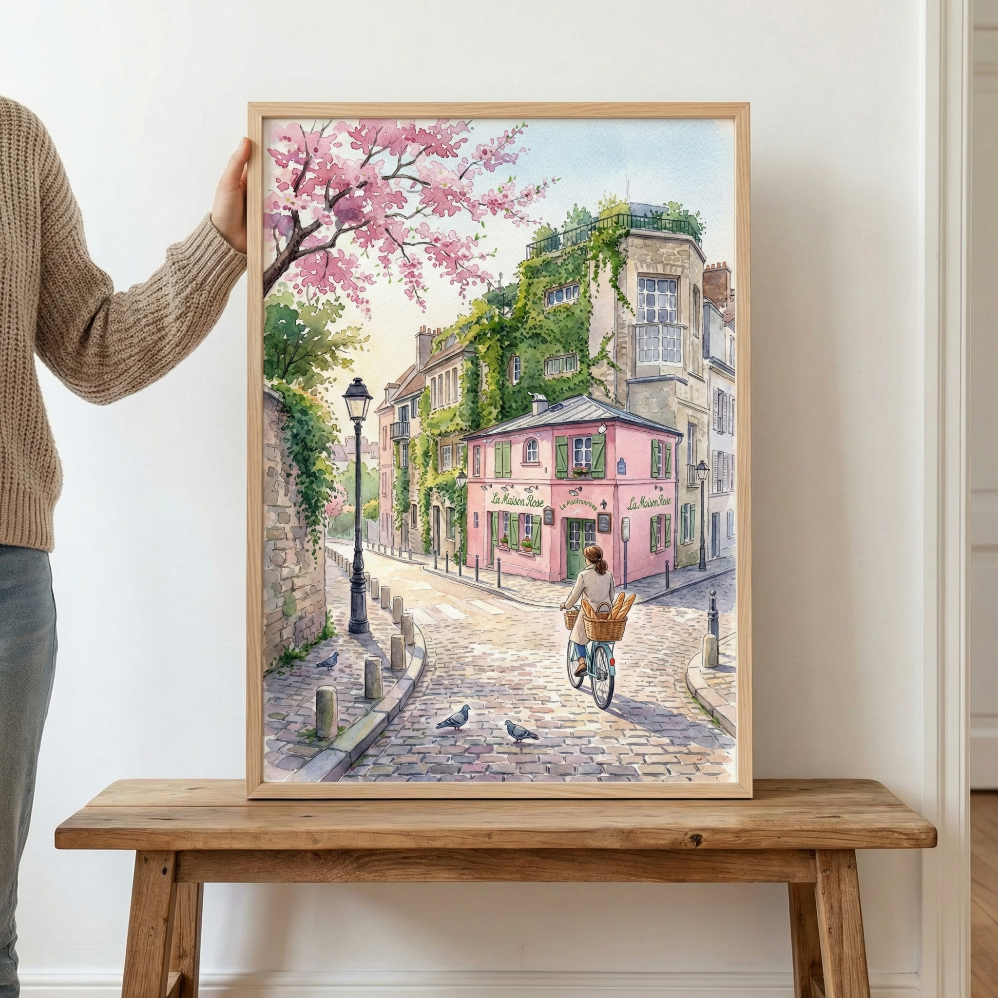 A person holds a framed watercolor painting depicting a romantic Parisian street corner. The artwork features the iconic pink building of La Maison Rose in Montmartre, with ivy-covered walls and classic architecture. A cyclist with a basket full of baguettes rides along a cobblestone street, while cherry blossoms bloom overhead and pigeons stroll nearby. The overall mood is peaceful, charming, and timeless.