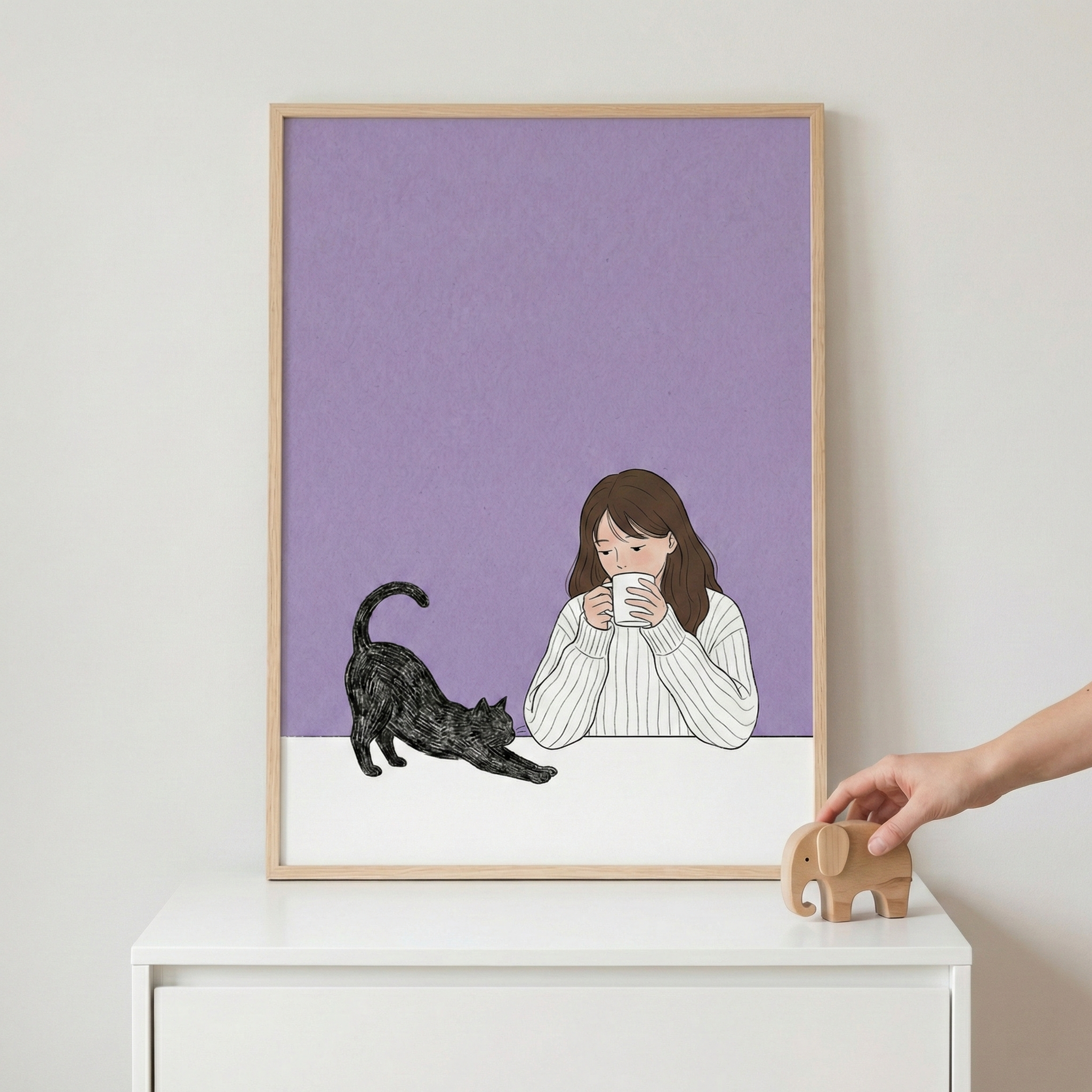 A light wood-framed illustration sits on a white dresser against a plain wall. The artwork depicts a woman with long brown hair in a white ribbed sweater holding a white mug to her face. Beside her, a black cat is shown stretching on a white surface against a light purple background. To the right of the frame, a person's hand reaches for a small, minimalist wooden elephant figurine.