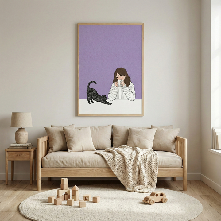 A framed illustration is centered on a light-colored wall, depicting a young woman with long brown hair sipping from a white mug. Beside her, a black cat is shown in a long stretch. The background of the illustration is a solid, matte purple. Below the art, there is a light-wood sofa with beige upholstery and several matching pillows. A thick, cream-colored knit throw is draped over the sofa.