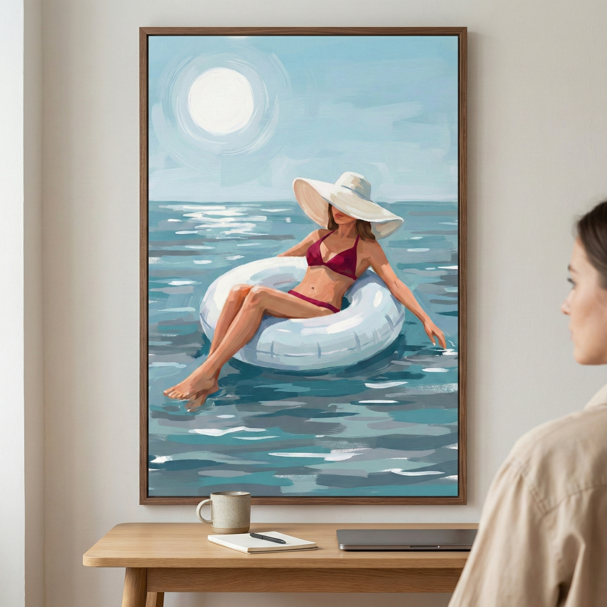 A vertical framed artwork hangs on a neutral wall above a wooden desk. The digital illustration depicts a woman in a maroon bikini and a large white sun hat, floating in a white inner tube on a vast, shimmering blue sea. A large, glowing sun hangs in the pale sky above her. The piece uses expressive, textured brushwork to create light and movement in the water. In the foreground, a person is partially visible, viewing the art. A mug, notebook, and laptop rest on the desk below.