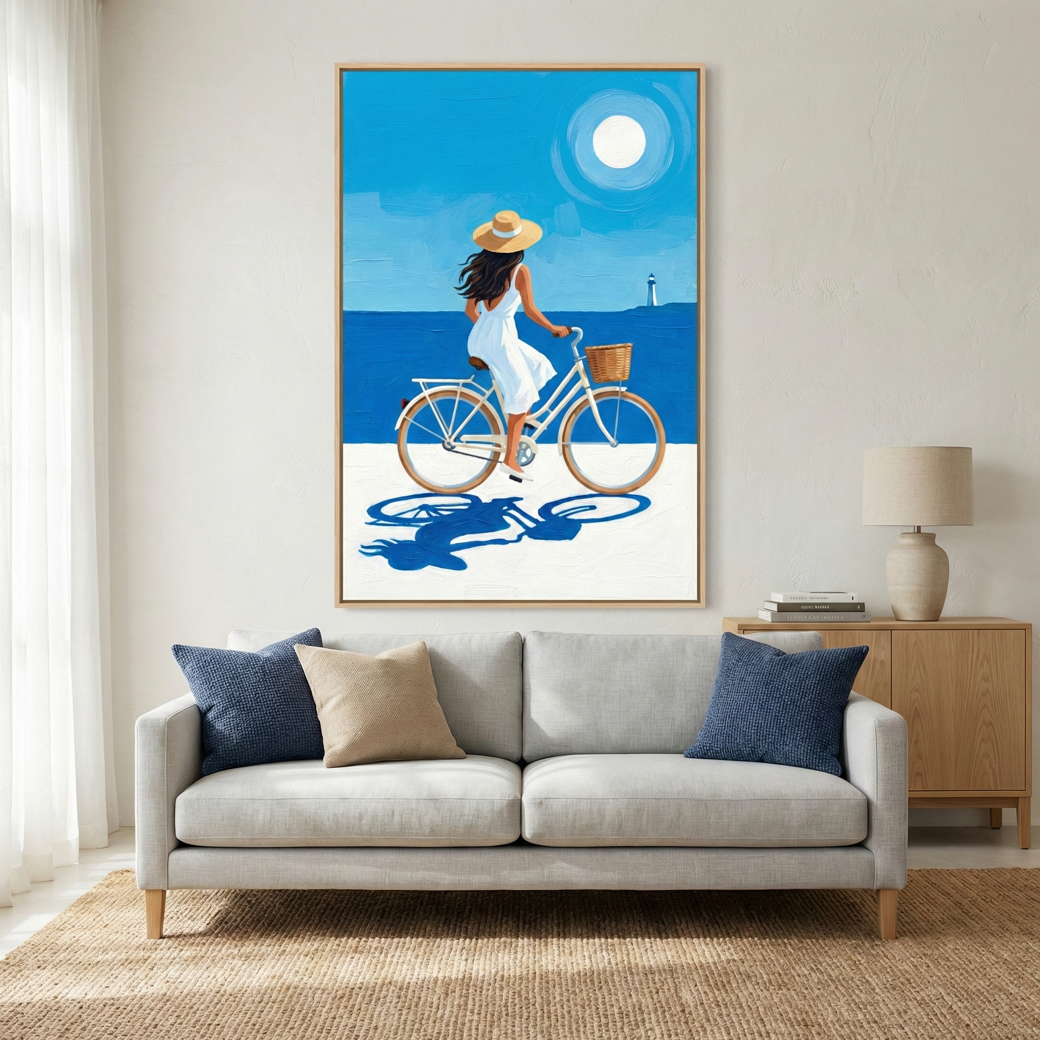 A large framed piece of wall art hangs above a modern light-gray sofa. The artwork is a stylized, flat-color digital illustration with a minimalist aesthetic, featuring bold, saturated blues and a clean, graphic layout reminiscent of classic travel posters.

Inside the frame, the scene depicts a woman with long, dark hair from behind, riding a vintage-style light-blue bicycle. She is wearing a simple white sundress and a wide-brimmed straw hat. The bicycle features a small woven basket on the front. 