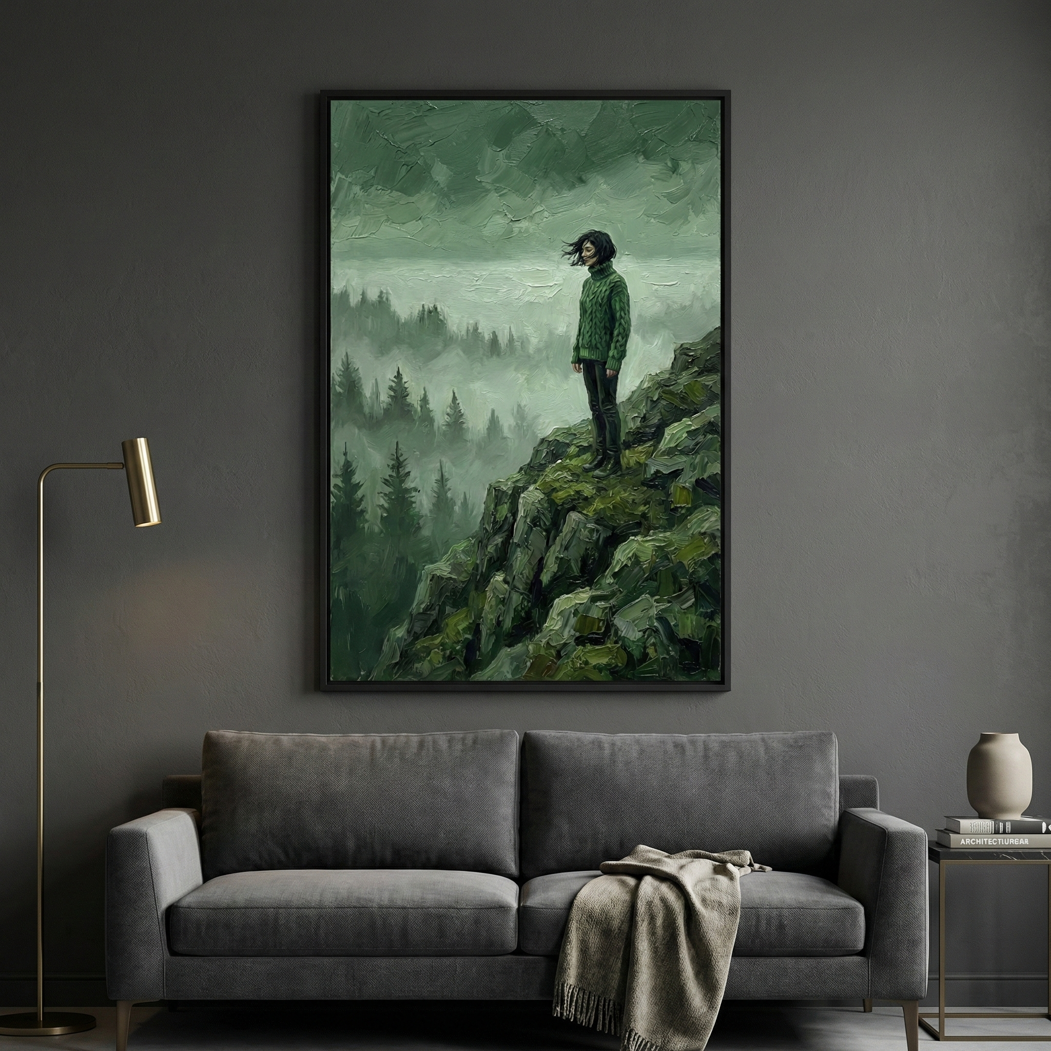 A moody interior with a dark gray wall and sofa, featuring a large framed painting of a woman standing at the edge of a forested cliff. Her black hair blows in the wind as she overlooks a foggy wilderness scene painted in rich green tones.