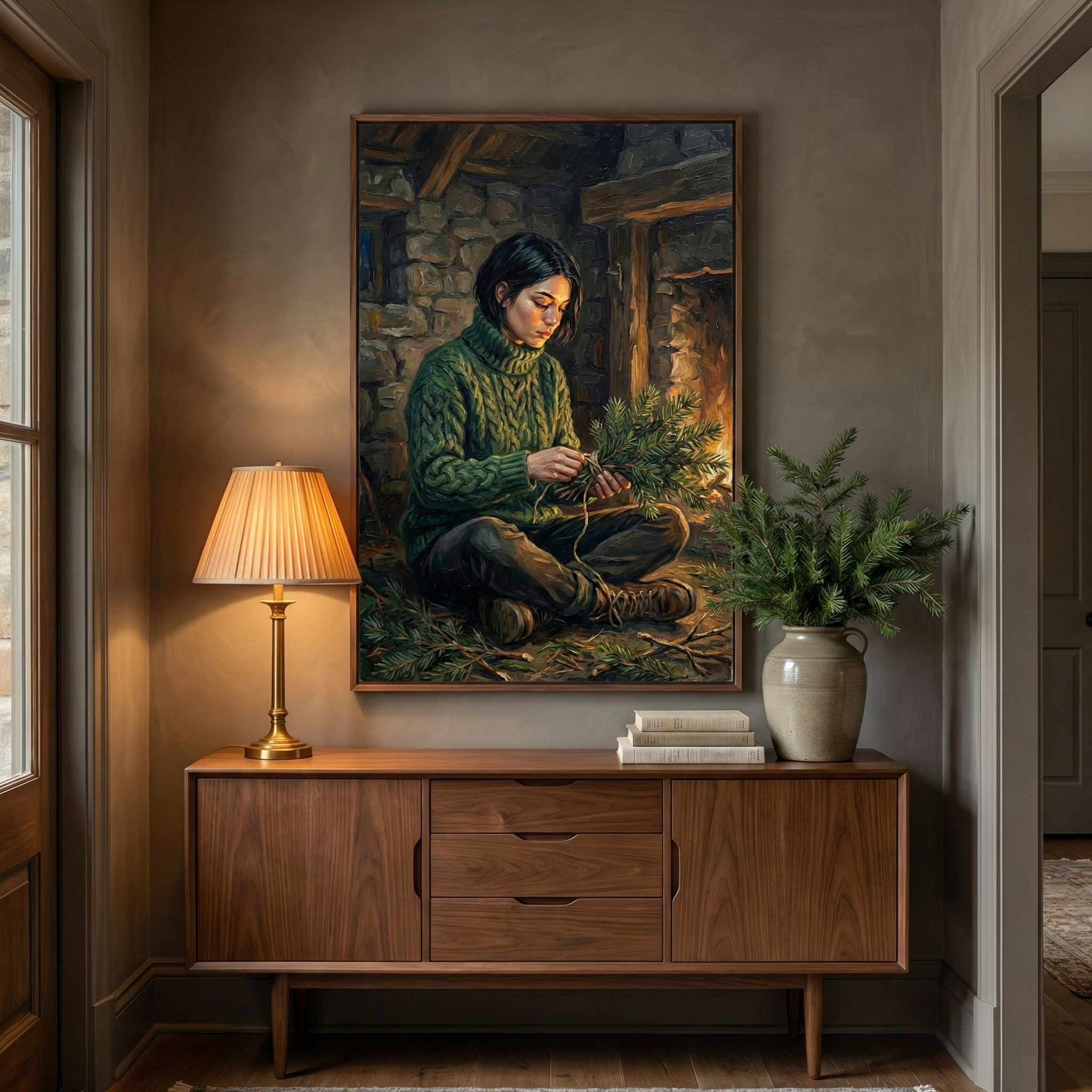 A cozy home interior with a mid-century modern wooden sideboard. Above hangs a large framed canvas illustrating a dark-haired woman in a green cable-knit sweater. She sits cross-legged in a rustic stone cabin, arranging a bundle of pine needles near a glowing fireplace. On the sideboard's left, a brass lamp with a pleated shade emits soft light. In the center is a small stack of books, and on the right, a tall beige vase holds lush evergreen boughs. The walls are a muted taupe.