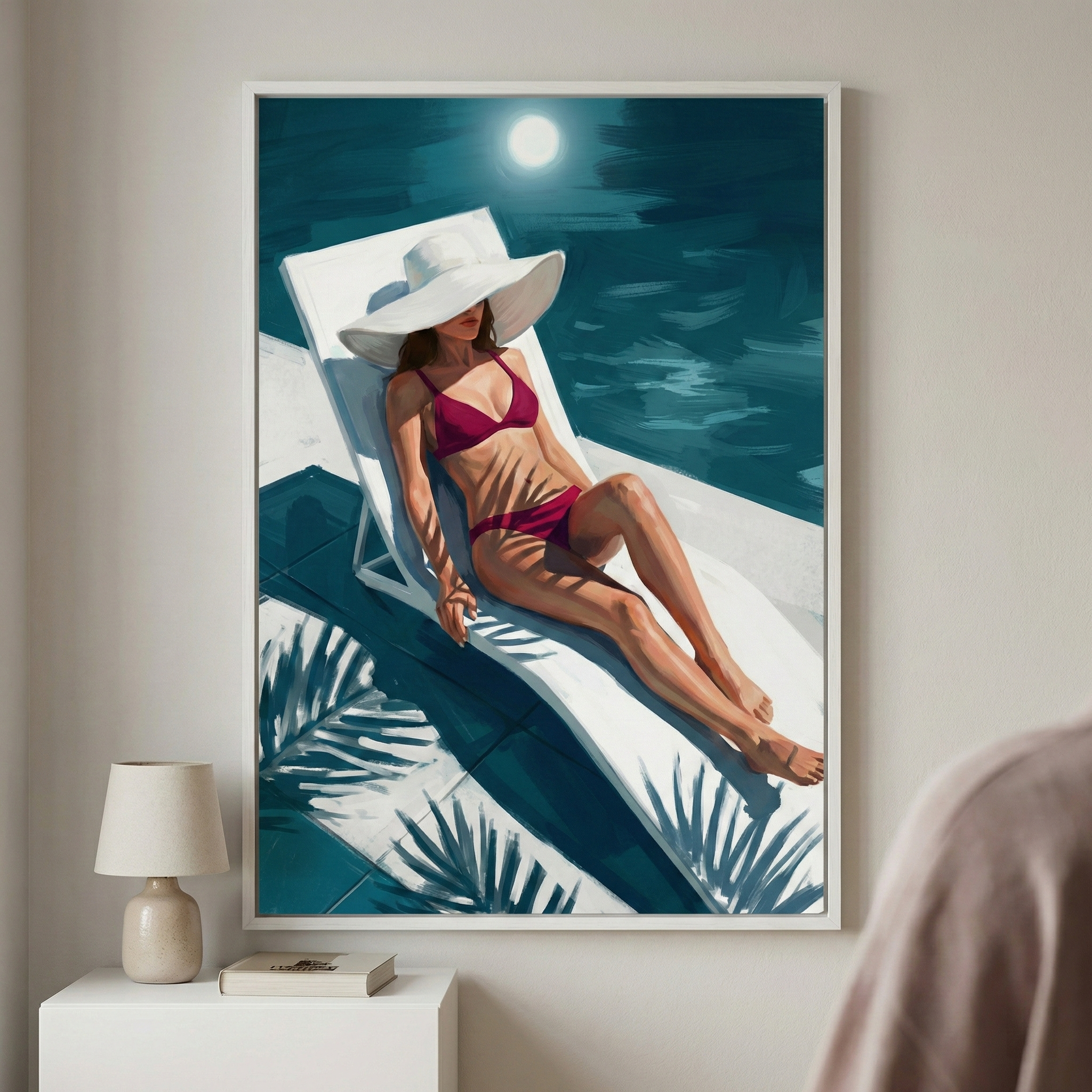 A framed piece of digital art hanging on a plain white wall above a small side table. The artwork depicts a woman in a maroon two-piece swimsuit and a wide-brimmed white hat, lounging on a white reclining chair. The setting is a moonlit night, with a bright moon in a dark teal sky reflected in the water of a pool behind her. Sharp shadows of palm leaves are cast over the woman and the lounge chair. 