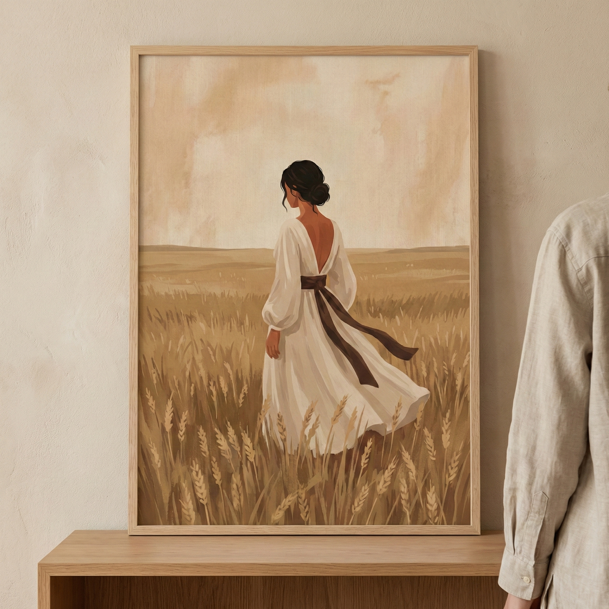 A piece of framed wall art shows a woman in a vast field of golden grain. Seen from behind, she looks toward the horizon under a soft, hazy sky. She wears a long white dress with full sleeves and a dark sash tied at her waist. The artwork uses a muted, earthy palette and soft textures. The frame is displayed on a wooden shelf against a neutral wall, with a person's linen sleeve visible to the right.