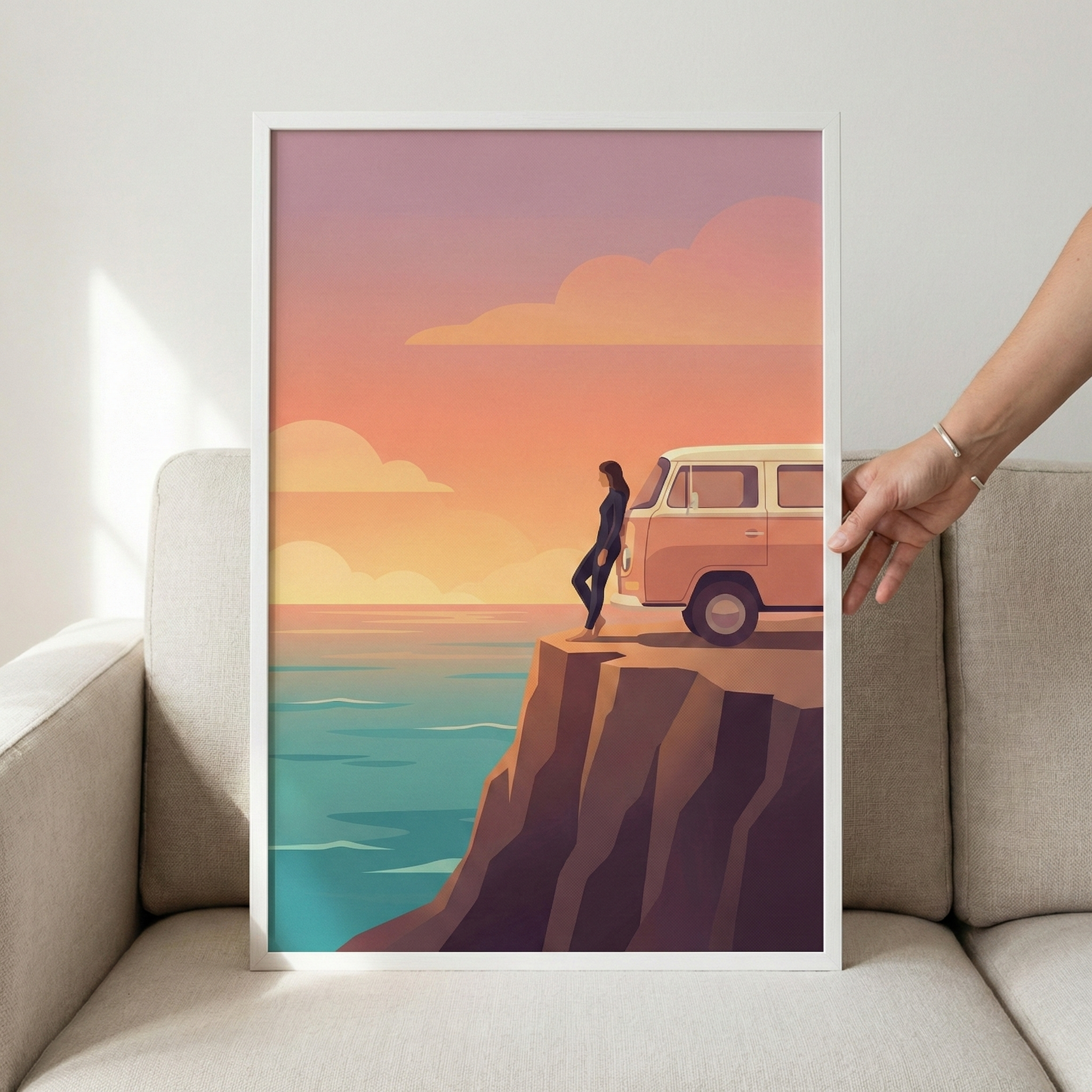 A minimalist digital illustration shows a woman leaning against a vintage van on the edge of a steep cliff. The scene overlooks a calm ocean under a warm, glowing sky at sunset, filled with soft pink and orange clouds. The water below is a mix of teal and blue hues. The cliff is rendered in dark purple and brown with simplified, angular shapes. The framed art piece is being held by a hand against the backdrop of a light-colored couch and wall.