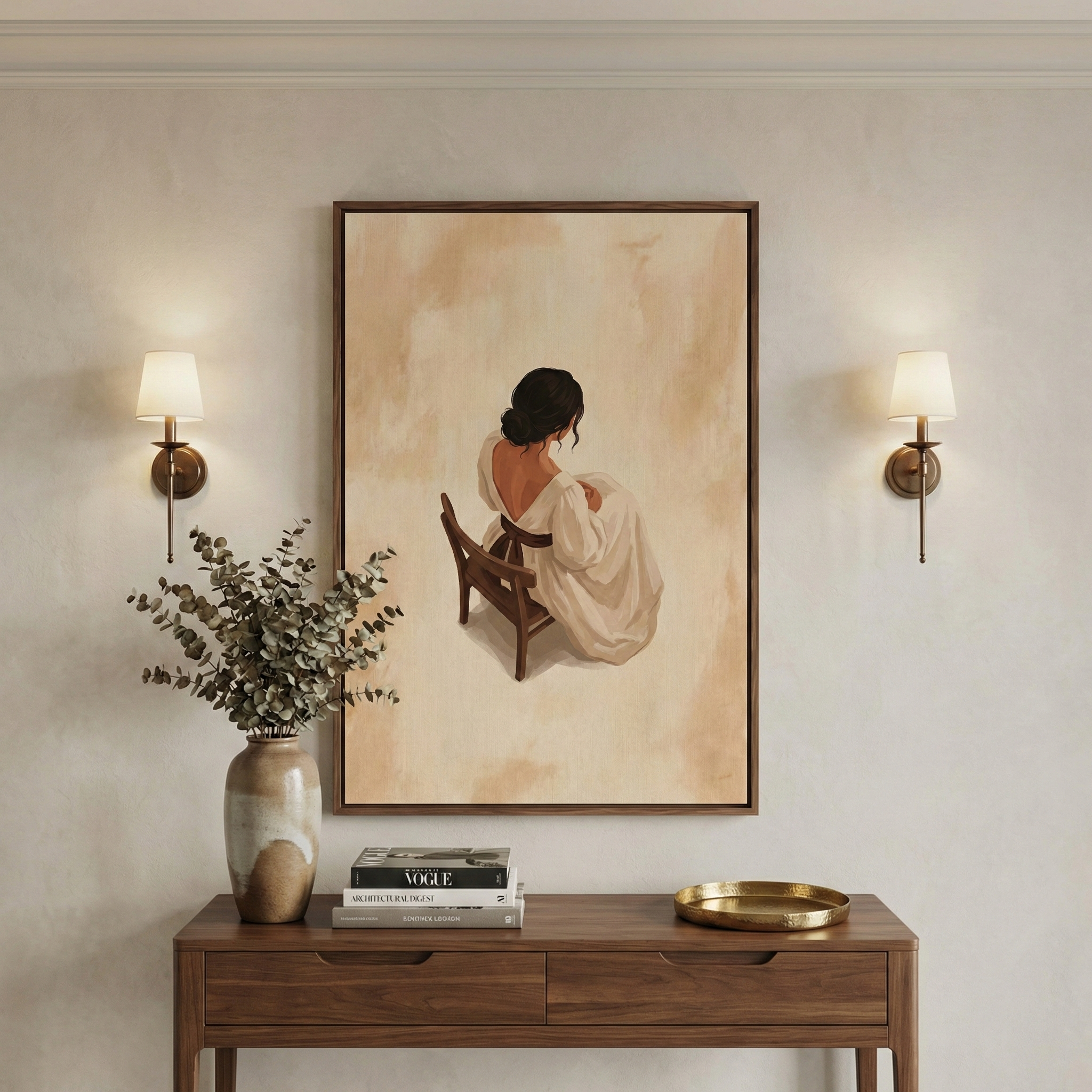 A balanced interior scene features a vertical, wood-framed artwork centered on a neutral wall. The art depicts the back of a woman with dark hair, wearing a white dress, seated on a small wooden chair. Flanking the art are two wall sconces with simple white shades. Below, a wooden console table holds a ceramic vase with green foliage, a small stack of books, and a shallow gold-colored tray. The entire setting is bathed in soft, warm light, creating a calm and minimalist aesthetic.