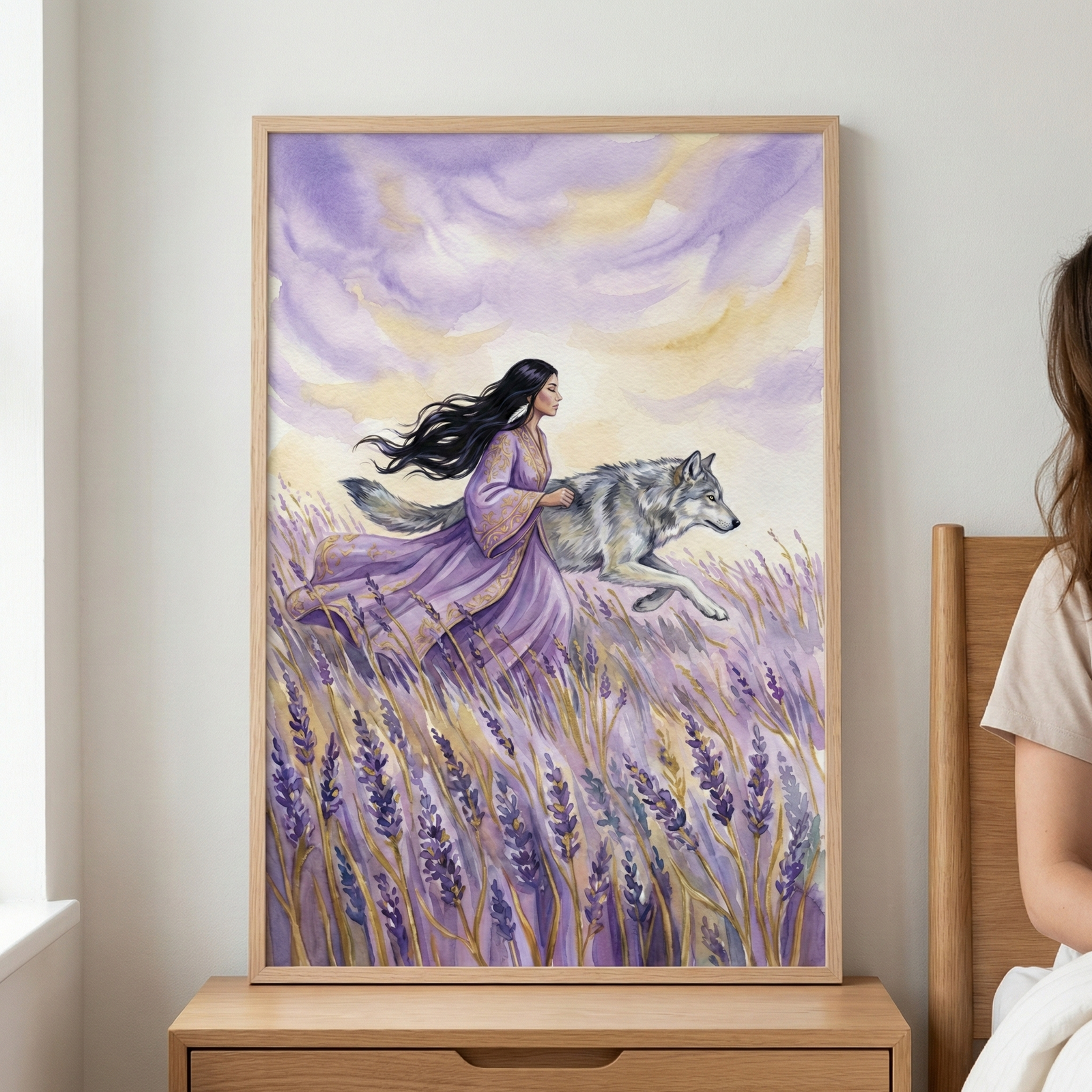 A framed watercolor illustration rests on a light wood nightstand. The artwork features a woman with long, flowing black hair and a regal purple gown with gold details, walking through a vibrant lavender field. A large gray wolf moves alongside her. The background shows an ethereal sky with soft purple and yellow clouds. The frame is a light-colored wood. To the right, a person's shoulder and arm are partially visible, sitting on the edge of a bed.