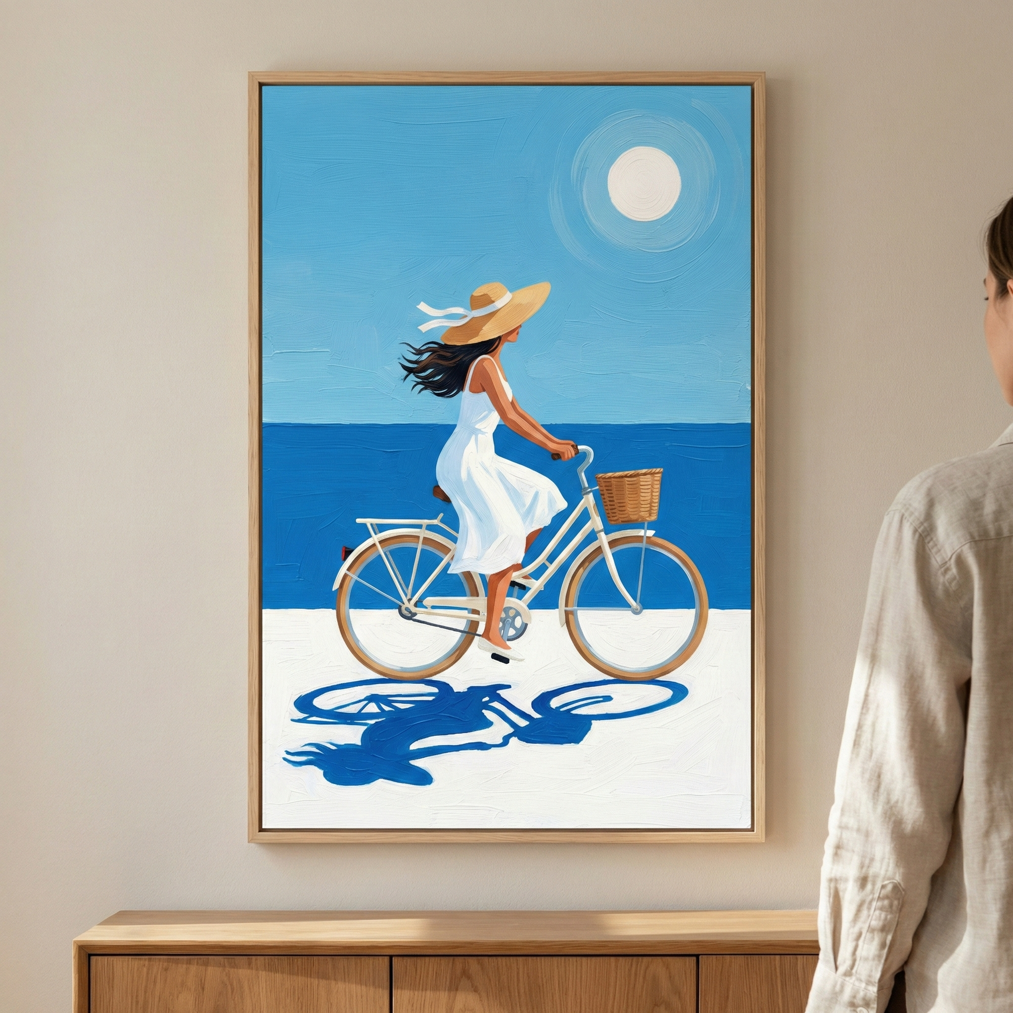 A framed vertical artwork hangs on a beige wall. It features a stylized illustration of a woman with dark hair wearing a white sleeveless dress and a wide-brimmed straw hat with a white ribbon. She is riding a light blue bicycle with a brown wicker basket and a rear rack. The scene is set on a white ground, with a flat deep blue sea and a light blue sky above. A bright, circular white sun shines in the upper sky. A dark blue, stylized shadow of the woman and the bike is cast on the ground. 