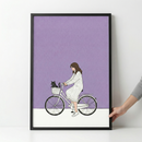 A minimalist illustration within a thin black frame shows a girl with long brown hair riding a white bicycle. She wears a long-sleeved, white and grey vertically striped dress. A small black cat sits tucked into the white basket at the front of her bike. The background consists of a large solid purple block over a clean white ground. The style is modern and clean with simple line work. A person's hand is visible on the right, holding the edge of the frame against a white wall.