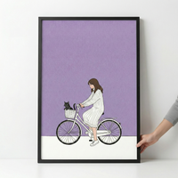 A minimalist illustration within a thin black frame shows a girl with long brown hair riding a white bicycle. She wears a long-sleeved, white and grey vertically striped dress. A small black cat sits tucked into the white basket at the front of her bike. The background consists of a large solid purple block over a clean white ground. The style is modern and clean with simple line work. A person's hand is visible on the right, holding the edge of the frame against a white wall.