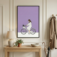 A vertical framed illustration hangs on a cream-colored wall above a light wood console table. The illustration depicts a girl with long brown hair, wearing a white ribbed dress, riding a light-colored bicycle. A small black cat sits in the bicycle's front basket. The background of the illustration is a solid light purple above a white floor line. On the wooden table below the frame, there is a neutral-toned lamp, a small green plant in a clay pot, and a shallow bowl. 