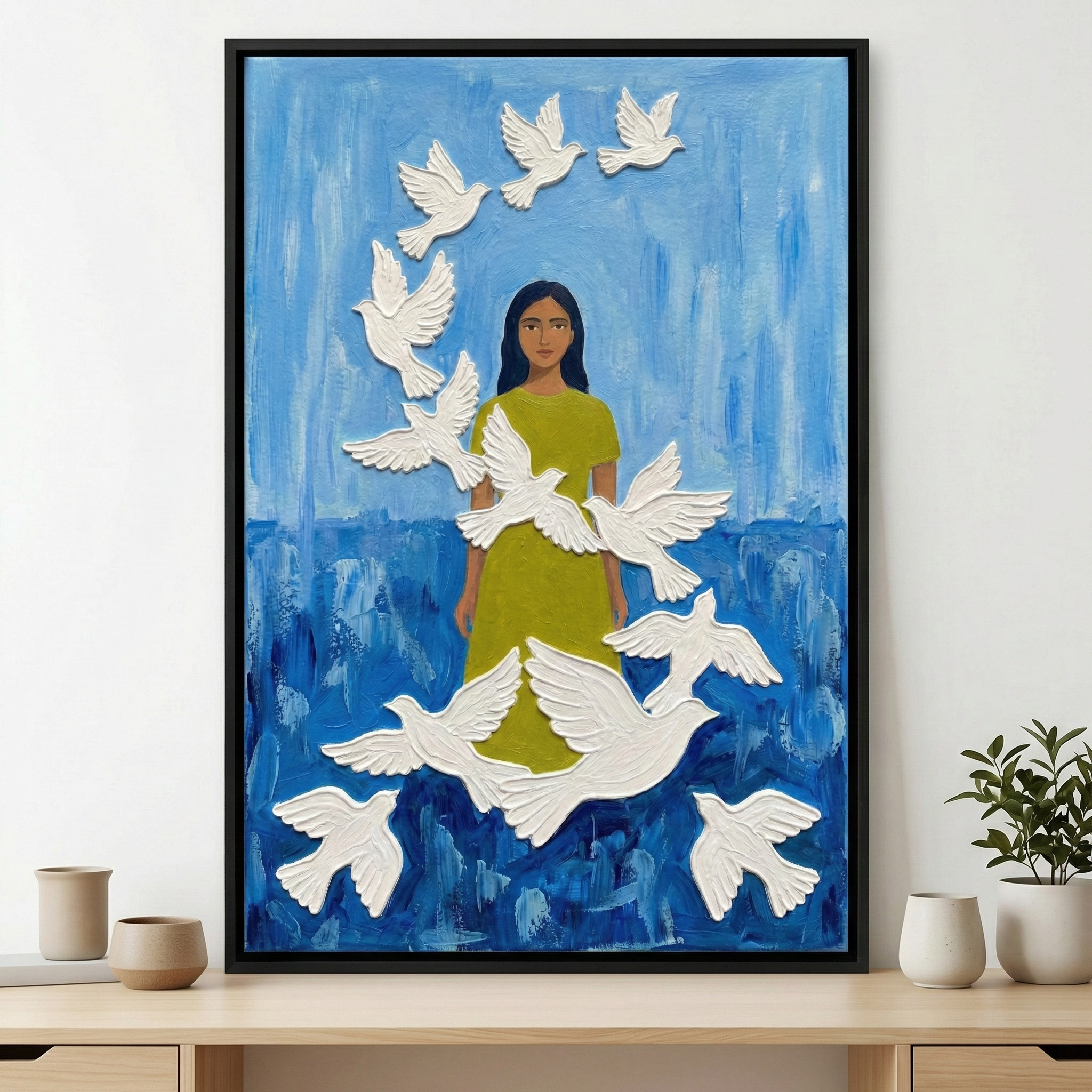 A framed artwork features a woman with dark hair and a lime-green dress, standing amidst a swirling flock of white birds. The birds are rendered with a thick, tactile texture, giving them a three-dimensional appearance. The background is a rich, expressive blue with visible, sweeping strokes of color. The piece is housed in a thin black frame and displayed on a light wooden table next to minimalist pottery and a small green plant, creating a serene and modern indoor setting.