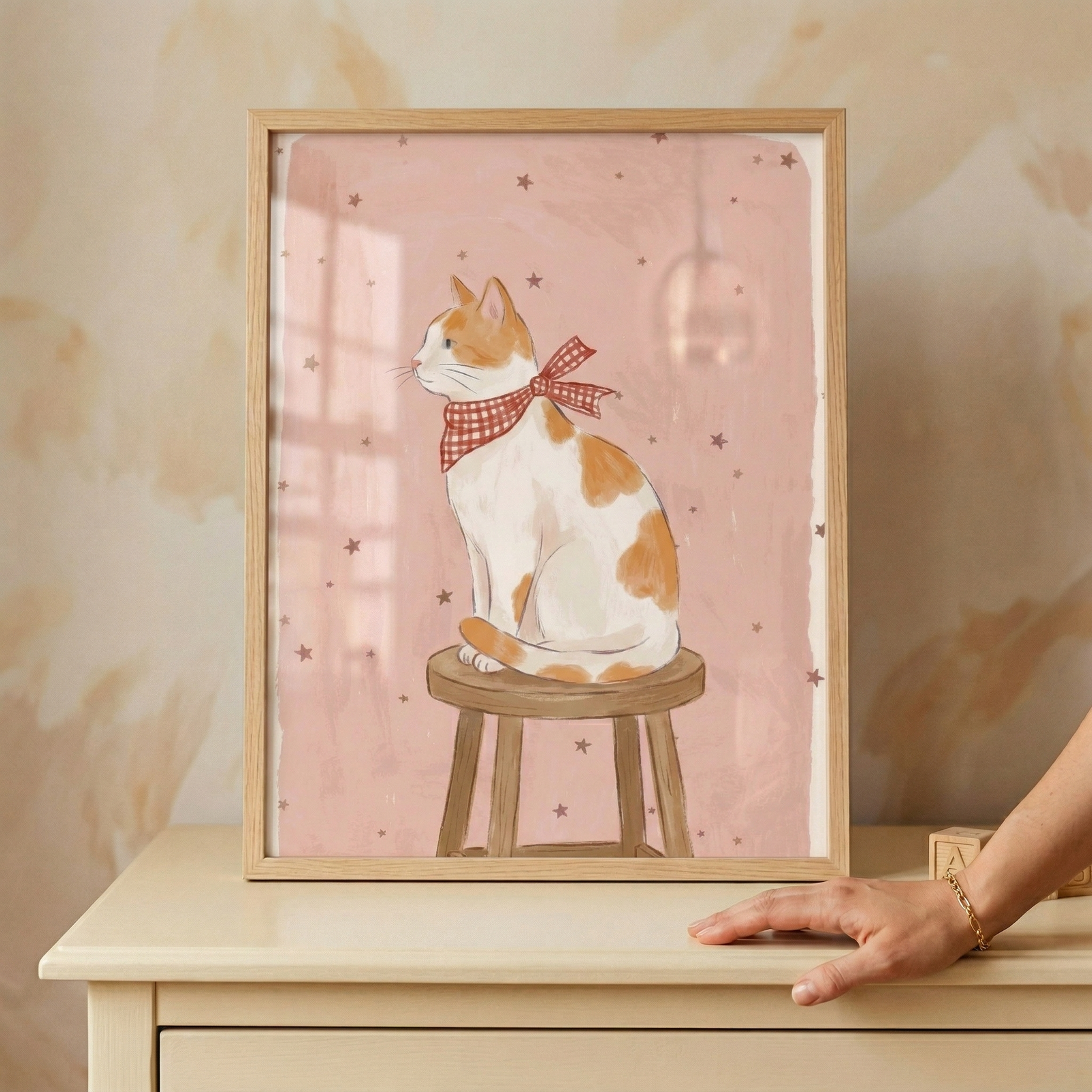 An illustration of a cat sits within a light wood frame, which is placed on top of a cream-colored dresser. The artwork features a white cat with orange patches sitting in profile on a simple, round-topped wooden stool. Around its neck is a red and white checkered bow. The cat is set against a soft pink background dotted with several small, brown stars.