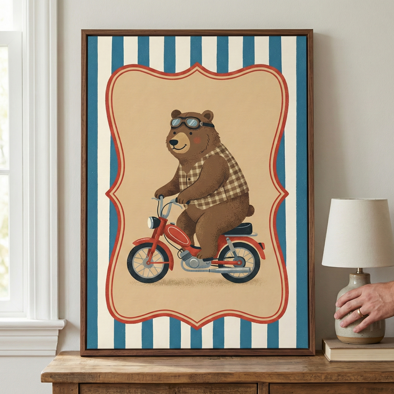 A framed illustration featuring a friendly brown bear riding a small red motorcycle. The bear is wearing a brown and cream checkered shirt and dark goggles pushed up on its forehead. The central scene is enclosed in a decorative, scalloped red border, which sits against a background of vertical blue and white stripes. The framed artwork is displayed on a wooden surface next to a small lamp and a book.