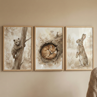 Three framed pieces of watercolor-style artwork hang in a row on a neutral beige wall. The artwork on the left features a brown bear cub climbing a tree trunk. The center piece depicts a fox curled up and sleeping inside a hollowed-out log. The artwork on the right shows a brown rabbit standing on its hind legs in a grassy field. Each piece is set in a light-colored wooden frame and uses a soft, earthy color palette. A small portion of a beige knit sweater is visible in the bottom right corner of the image.