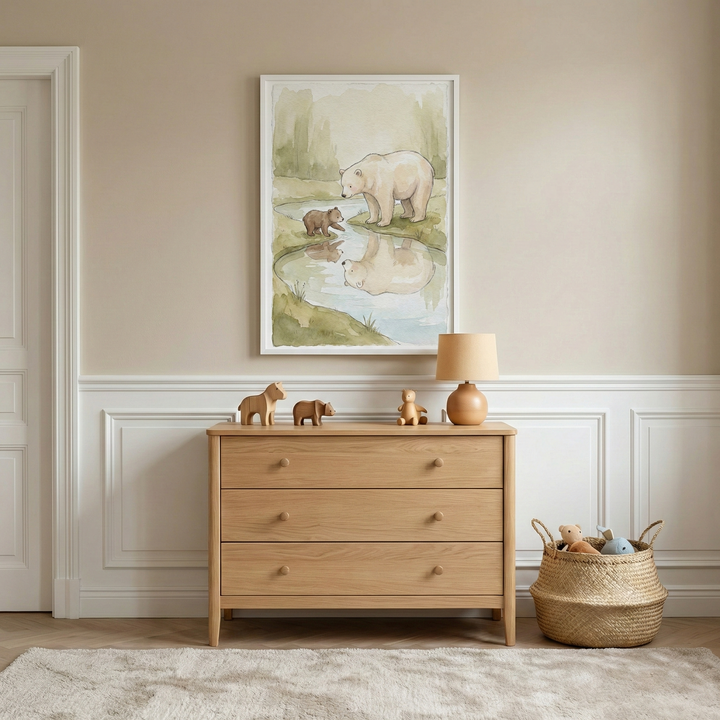 A peaceful nursery scene showcases a light wood three-drawer chest against a beige wall with elegant white wainscoting. On the dresser sit a small wooden horse, a bear figurine, a tiny figure, and a lamp with a wooden base and beige shade. Hanging above is a framed watercolor-style artwork of a large white bear and a brown cub reflected in a stream. To the right, a woven basket holds plush toys. A soft white rug lies on the floor, and a white paneled door is visible to the left.