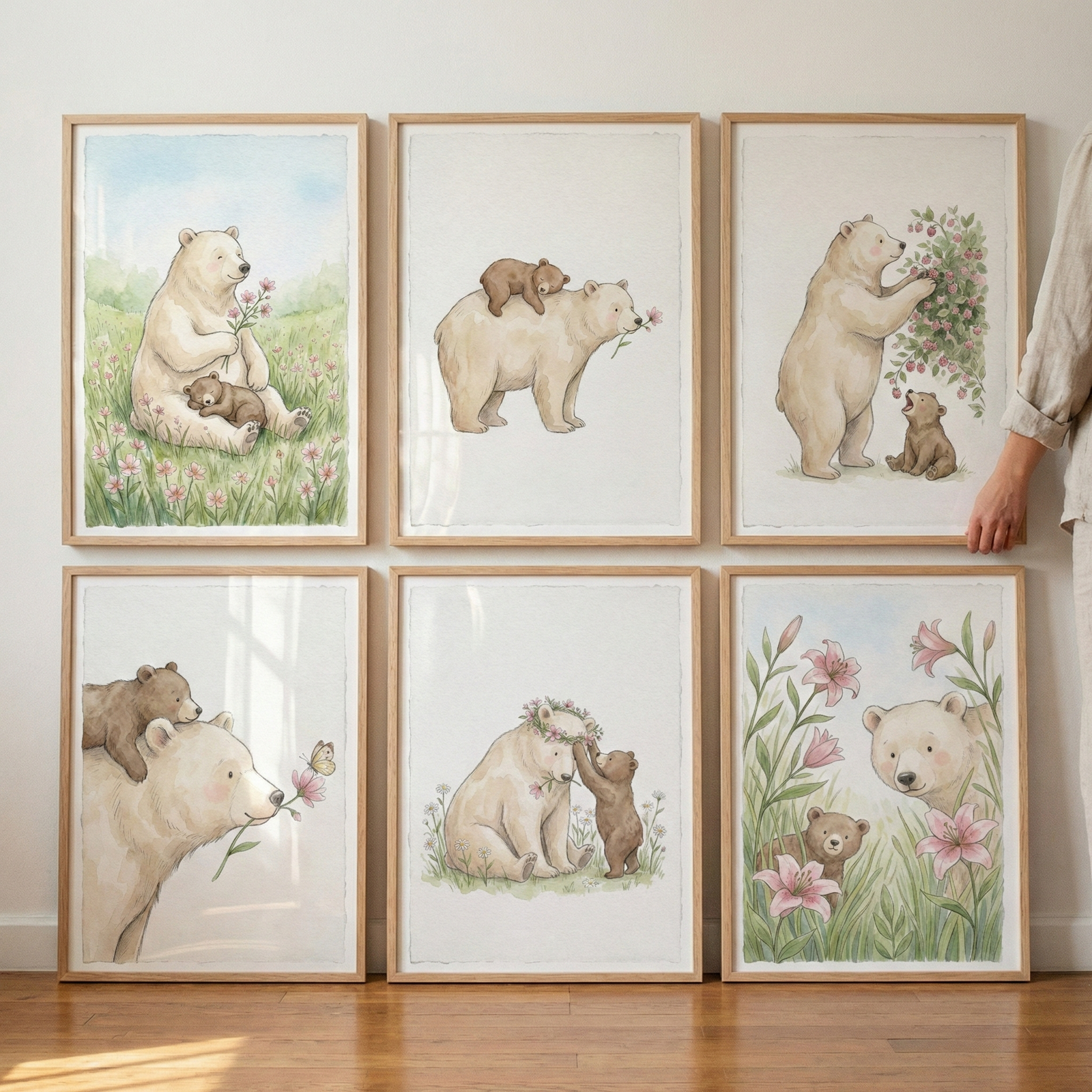 Six framed artworks in a watercolor style depict a large cream-colored bear and a brown cub. The top row shows the pair in a meadow, the cub riding on the bear's back, and picking berries. The bottom row shows them with a butterfly, a floral crown, and peeking through tall lilies. All pieces are in light wood frames, arranged in two rows of three against a white wall. A hand is visible on the right, adjusting a frame. The illustrations use soft, gentle tones.