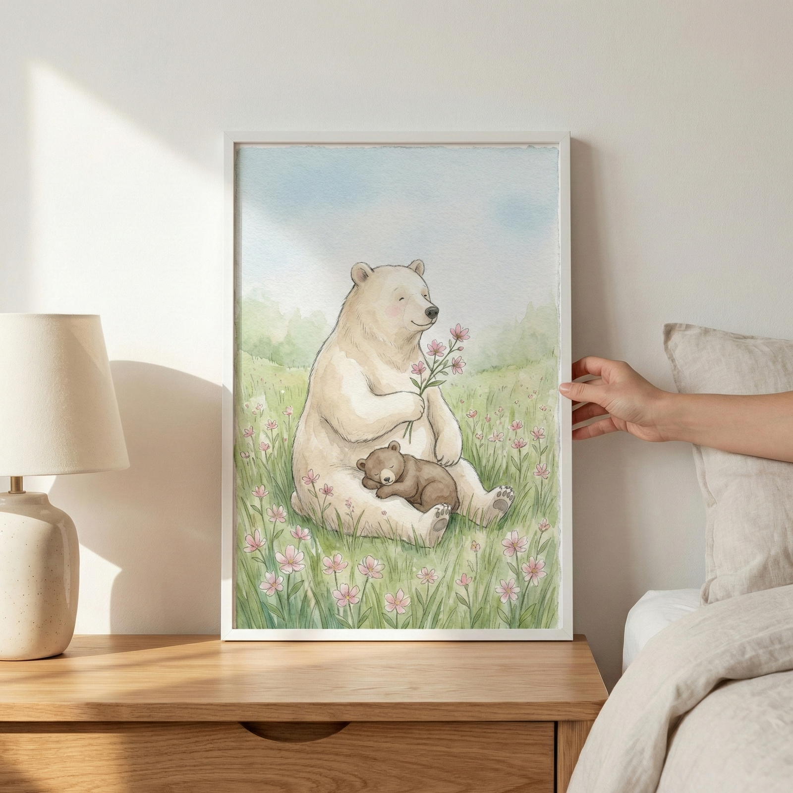 A framed watercolor illustration depicts a large, off-white bear sitting in a grassy meadow filled with pink wildflowers. The bear holds a small bouquet of pink blossoms while a tiny brown cub sleeps soundly against its belly. The background features rolling green hills and a soft blue sky. The piece is in a white frame, held by a hand against a wall next to a bed. A lamp sits on a wooden nightstand to the left, with warm light casting soft shadows.