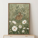 A framed illustration depicts a small, brown rabbit nestled in a lush meadow. Surrounded by tall, sage-green grasses and delicate leafy stems, the rabbit sits among various wildflowers. Large white blossoms with yellow centers are prominent in the foreground, while smaller, dainty white flowers and seed heads are scattered throughout the background. The artwork features soft textures and a muted color palette of earthy greens and browns. It is displayed in a thin wooden frame.