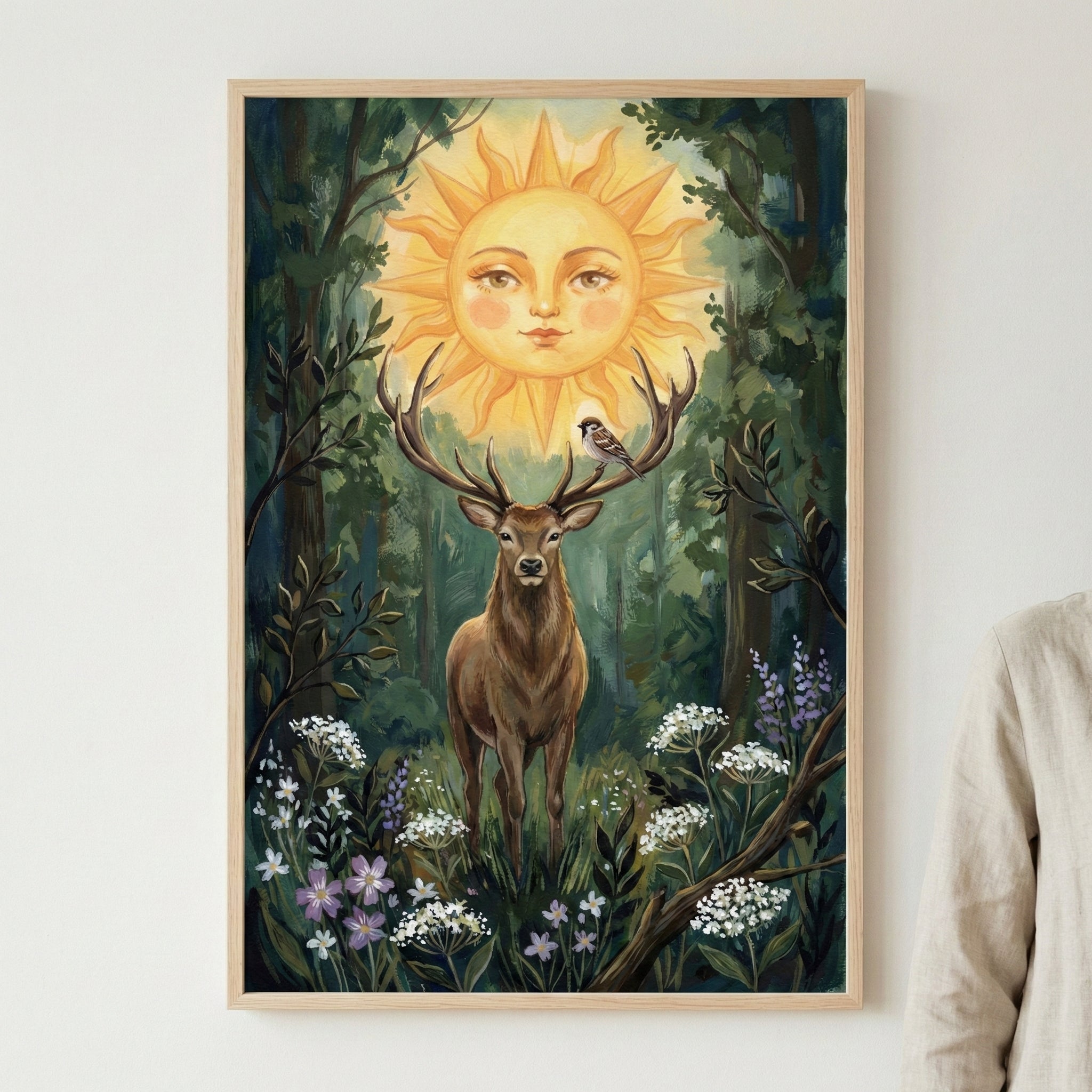 A vertical piece of wall art depicting a brown deer with large antlers in a deep green forest. Positioned between the antlers is a glowing sun with a calm human face. A small sparrow-like bird is perched on one of the antlers. The foreground features clusters of white and lavender wildflowers amongst dense foliage. The artwork is encased in a simple, light-toned wooden frame and displayed on a neutral wall.