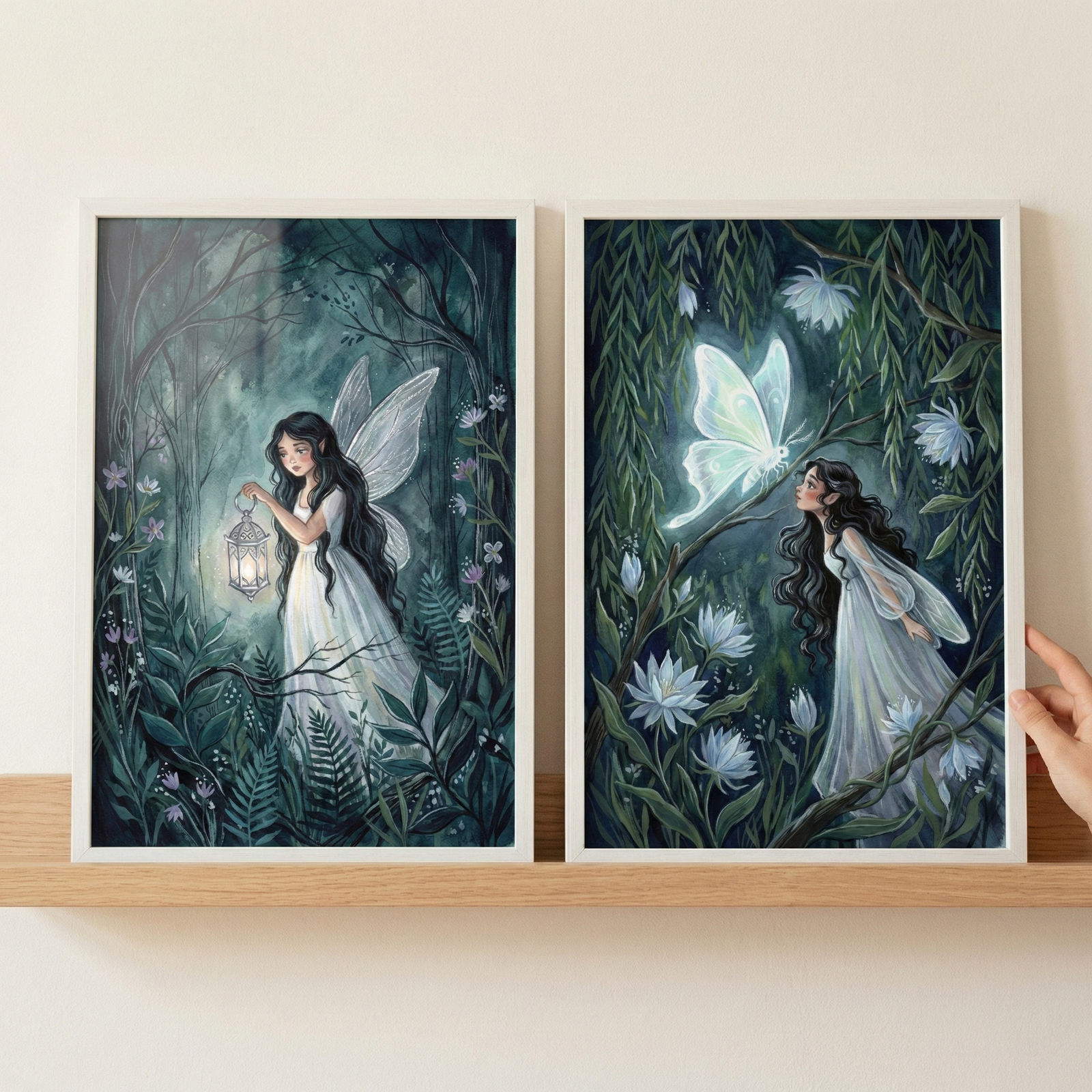 Two framed vertical art pieces sit on a light wood shelf against an off-white wall. The left illustration features a woman with long dark hair and delicate wings in a moonlit forest, holding a glowing lantern. The right piece shows a woman in a white dress looking up at a large, luminous, ethereal butterfly amidst vines and pale flowers. Both scenes use deep greens and teals with soft, glowing white highlights. A hand is visible on the far right, touching the edge of the frame.