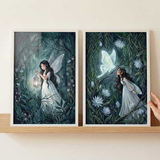 Two framed vertical art pieces sit on a light wood shelf against an off-white wall. The left illustration features a woman with long dark hair and delicate wings in a moonlit forest, holding a glowing lantern. The right piece shows a woman in a white dress looking up at a large, luminous, ethereal butterfly amidst vines and pale flowers. Both scenes use deep greens and teals with soft, glowing white highlights. A hand is visible on the far right, touching the edge of the frame.