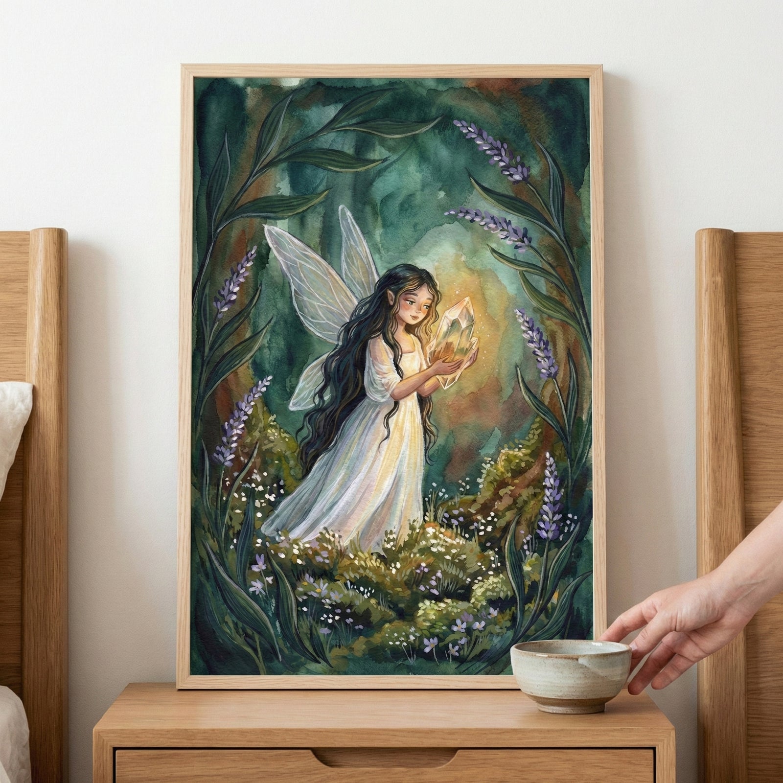 A framed fantasy illustration of a fairy with long dark hair and translucent wings, standing in a mystical forest. She is wearing a flowing white dress and holding a glowing crystal that illuminates her face. The scene is framed by tall, arcing green foliage and stalks of purple flowers. The ground is covered in moss and tiny wildflowers. The artwork is displayed in a light wood frame on a wooden nightstand, where a hand reaches for a small ceramic bowl.