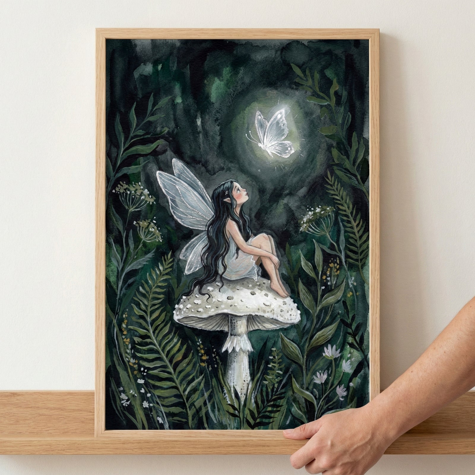 An illustration in a wooden frame depicts a small, winged figure with long dark hair sitting on top of a large, white-spotted mushroom. The figure looks upward at a glowing white butterfly in a dark, dense forest filled with ferns and delicate white flowers. The artwork features deep shades of green and grey. A hand is visible at the bottom right corner, holding the frame as it sits on a wooden shelf.