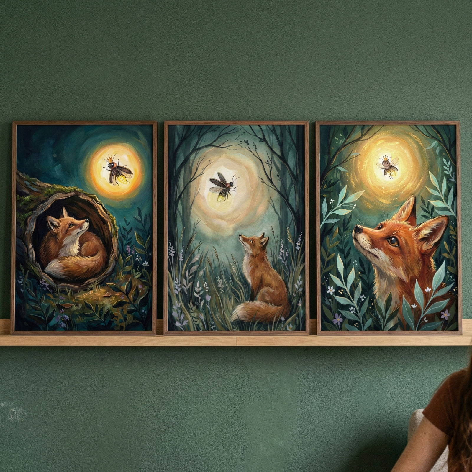 Three framed illustrations are displayed on a wooden shelf against a dark green wall. Each artwork depicts a red fox and a glowing winged insect in a forest at night. In the left frame, a fox curls in a hollow log looking up. The center frame shows a fox in a clearing gazing at a large orb of light. The right frame features a close-up of the fox's face among leaves. The style is whimsical with soft, warm light, creating a magical atmosphere.