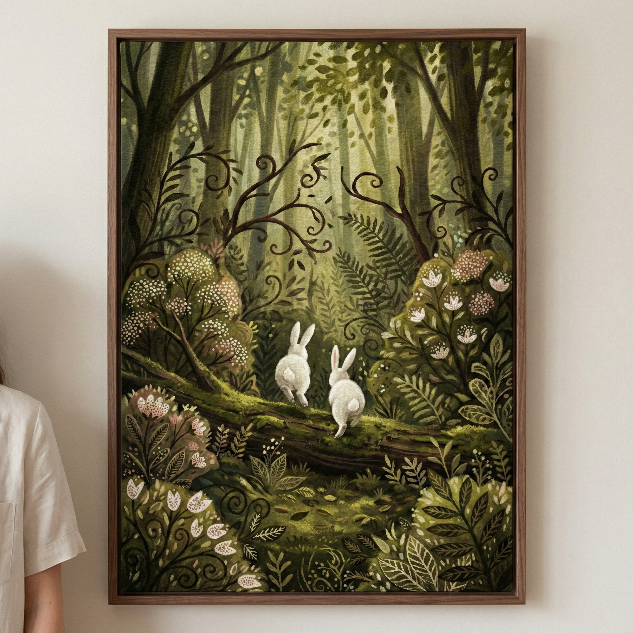 A framed vertical artwork hangs on a wall. The illustration depicts a whimsical, lush forest scene. Two small white rabbits hop away along a mossy fallen log toward a bright, sunlit clearing. The forest is filled with intricate botanical details: ferns, stylized flowers in white and pink, and dark, curling branches. Soft light filters through tall trees in the background, creating a magical glow. The color palette features deep greens. A person in a white shirt is partially visible on the left for scale.