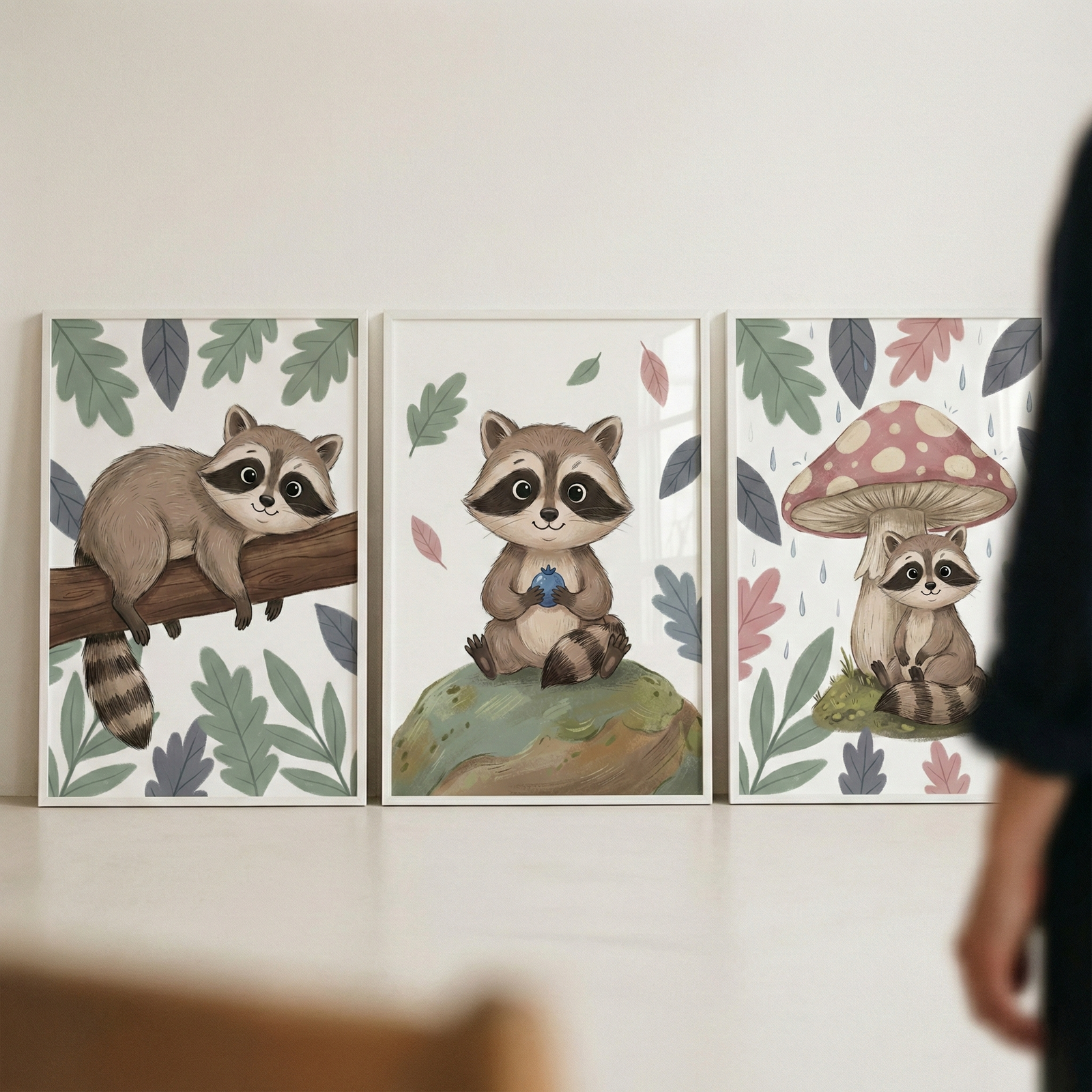 Three white-framed illustrations of a cute, cartoon raccoon are displayed side-by-side against a plain wall. The left illustration features the raccoon lounging on a thick tree branch, surrounded by green and purple leaves. In the middle, the raccoon sits on a mossy hill, holding a small blue berry while colorful leaves drift around it. The right illustration shows a small raccoon sitting under a large, red-and-white spotted mushroom during a gentle rain. The artwork has a soft, whimsical style.
