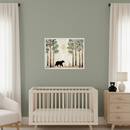 Modern nursery with sage green walls. A framed art print showing a black bear walking through a forest with a golden moon hangs above a light wood crib. A light wood dresser with a lamp and a black teddy bear is to the right, and a white armchair is to the left. White curtains are on either side of the room.