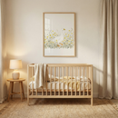 A serene and minimalist nursery setting featuring a light-toned wooden crib. Layered across the mattress are textured blankets in shades of cream and soft yellow. Directly above the crib hangs a framed artwork in a matching wooden frame, depicting a white goose wandering through tall grass and delicate yellow wildflowers. To the left, a small wooden side table holds a ceramic lamp with a fabric shade. A large, natural-fiber woven rug covers the floor beneath the furniture.
