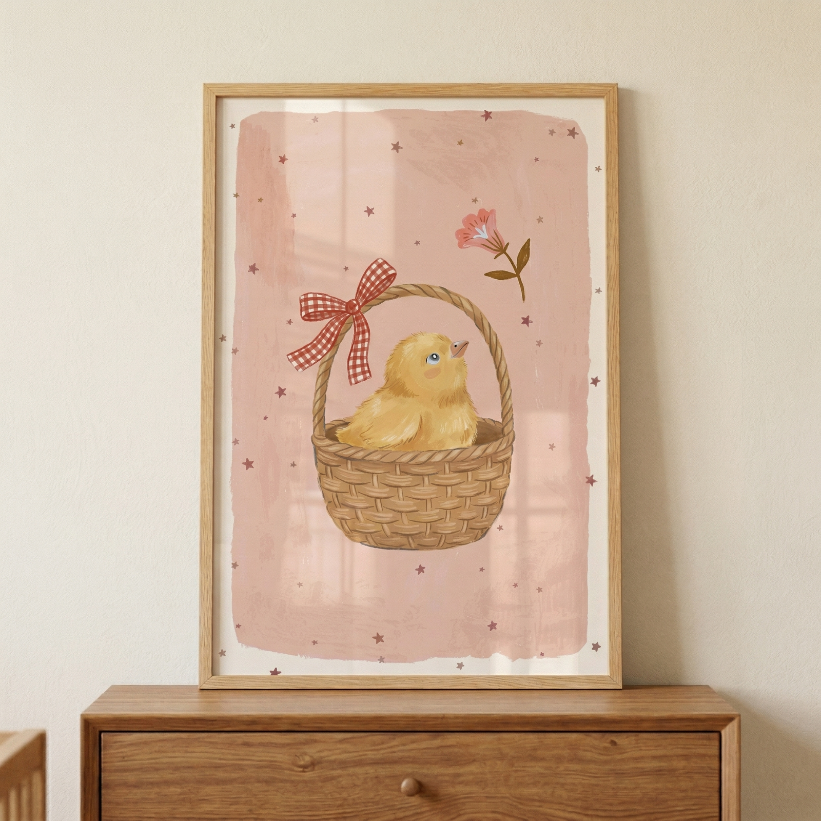 A framed vertical artwork sits on a wooden chest of drawers. The illustration inside the light wood frame features a small, fluffy yellow chick nestled inside a woven brown basket. A red and white checkered bow is tied to the basket's handle. The chick has a small orange beak and is looking upward. The background of the illustration is a soft, dusty pink with a textured appearance, accented by small dark stars scattered throughout. 