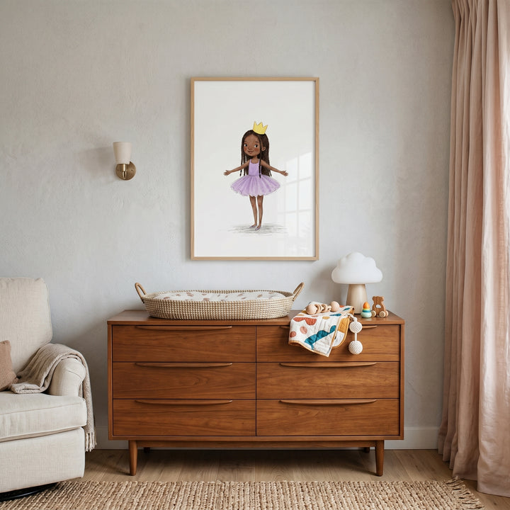 Framed yellow crown ballerina girl wall art hangs above a wooden dresser with a changing basket in a neutral nursery.