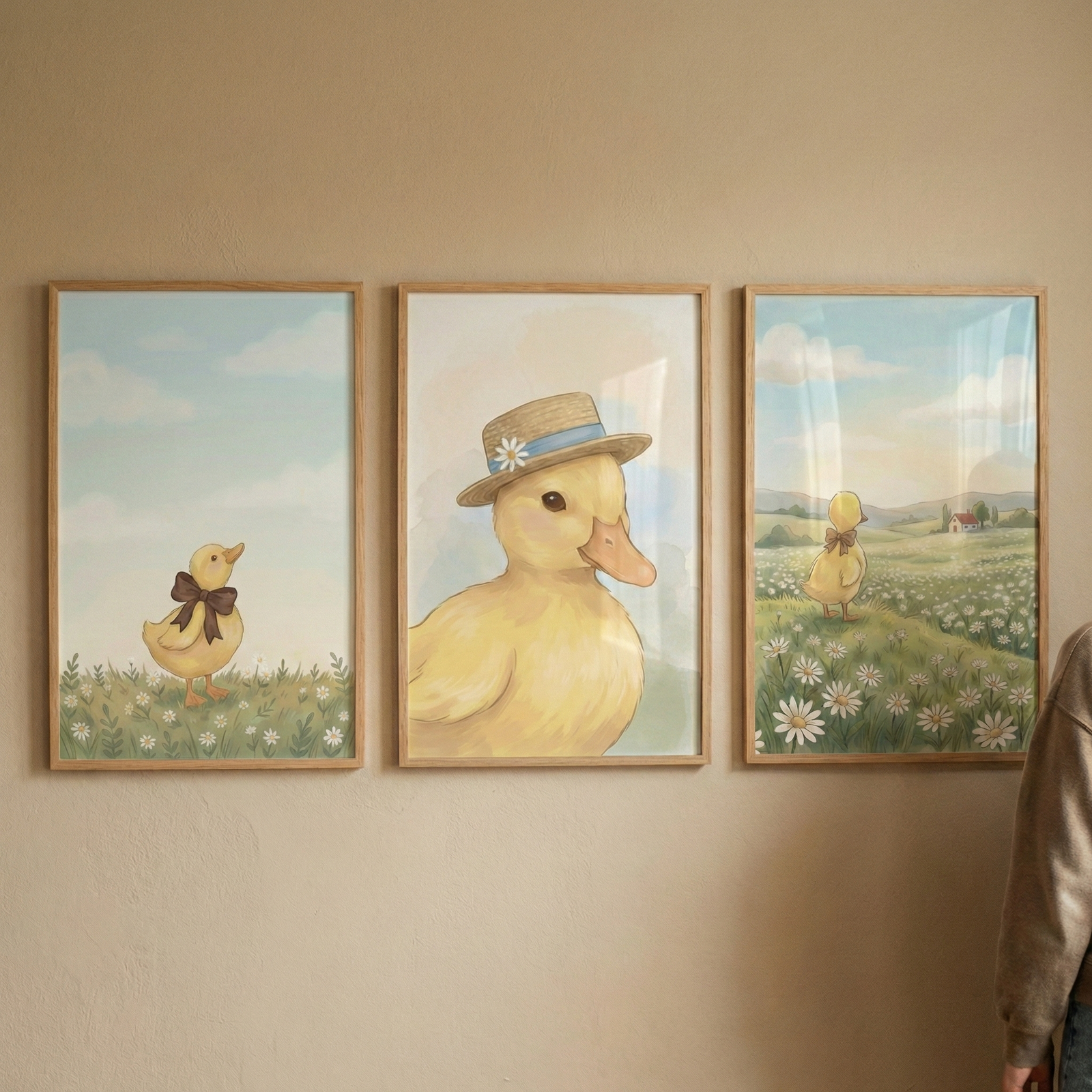 Three light wood frames hang on a neutral wall, each containing a whimsical illustration of a yellow duckling. The left artwork depicts the duckling with a brown bow in a grassy field. The center frame shows a close-up of the duckling wearing a straw hat. The right frame features the duckling walking away through a vast field of daisies towards a small red house under a soft, sunny sky. The style is gentle and soft-toned. A person's arm and shoulder are visible on the right edge.